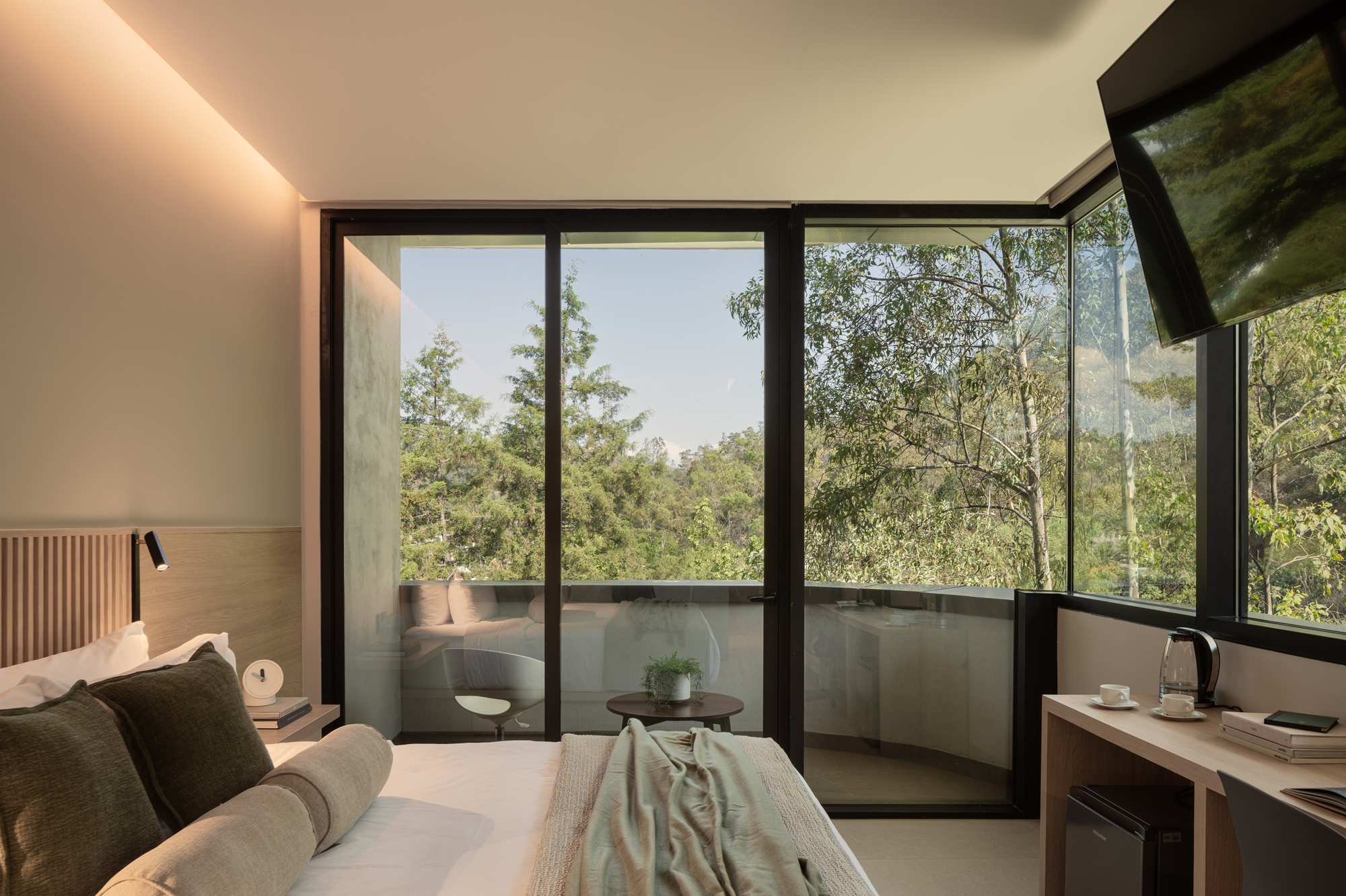 Hotel room corner with sliding glass doors opening to a balcony facing tree-covered hills