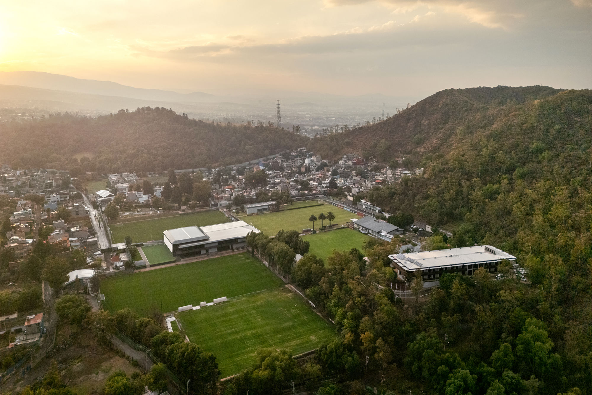 Aerial view of training fields and low pavilion structures nestled between forested hillsides at dusk