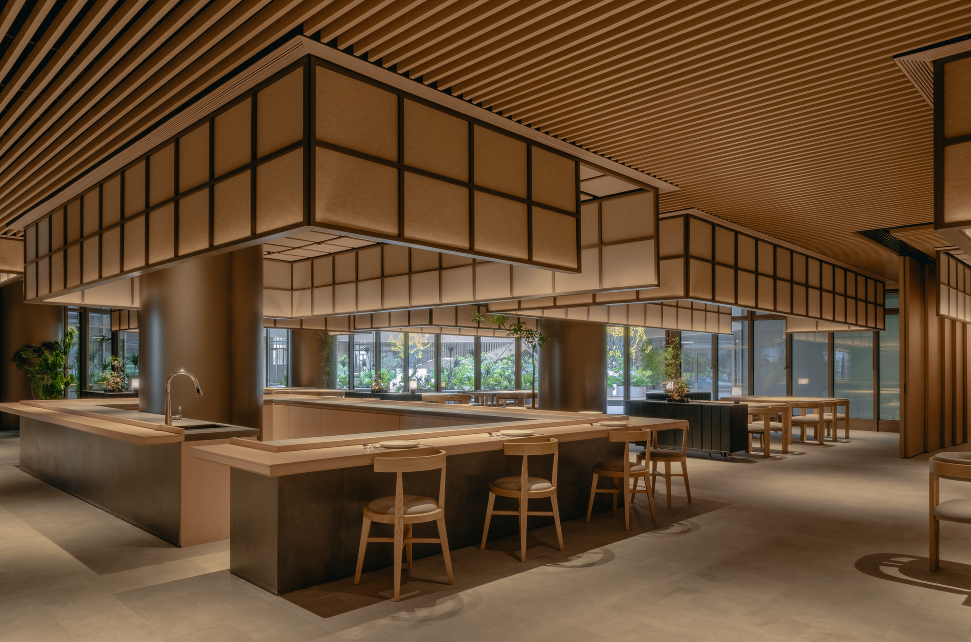 Long communal table with timber stools beneath suspended gridded panels and glazed courtyard beyond