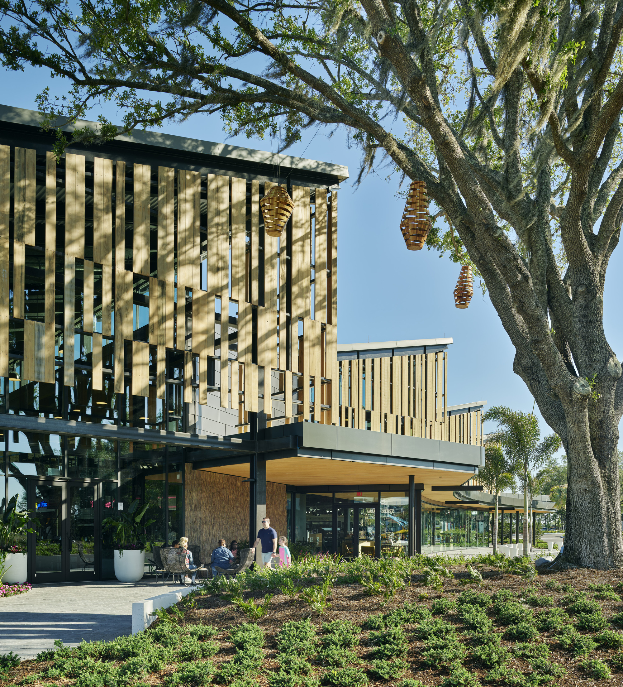 Two-story pavilion with vertical timber louvers casting shadows through overhanging oak branches with woven seed pods