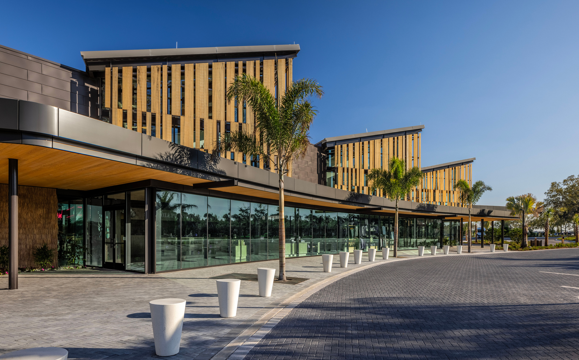 Glass-walled ground floor with white bollards lining a paved plaza beneath staggered timber-clad upper volumes
