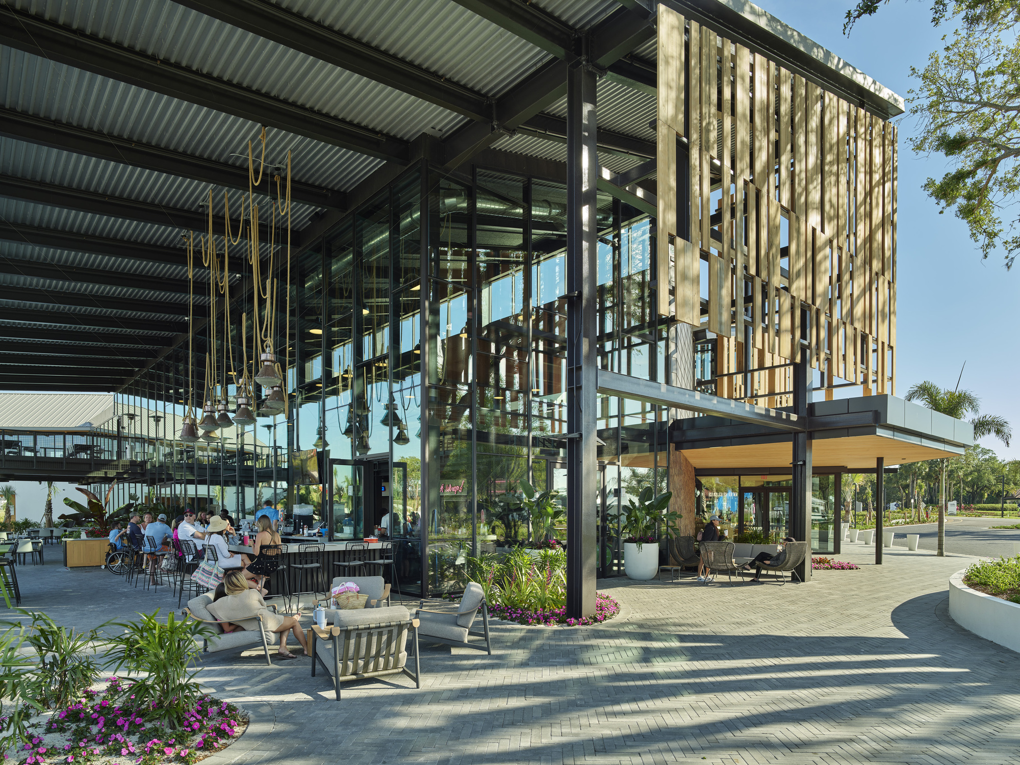 Covered outdoor dining terrace with steel columns, corrugated metal roof and vertical timber screen under blue sky