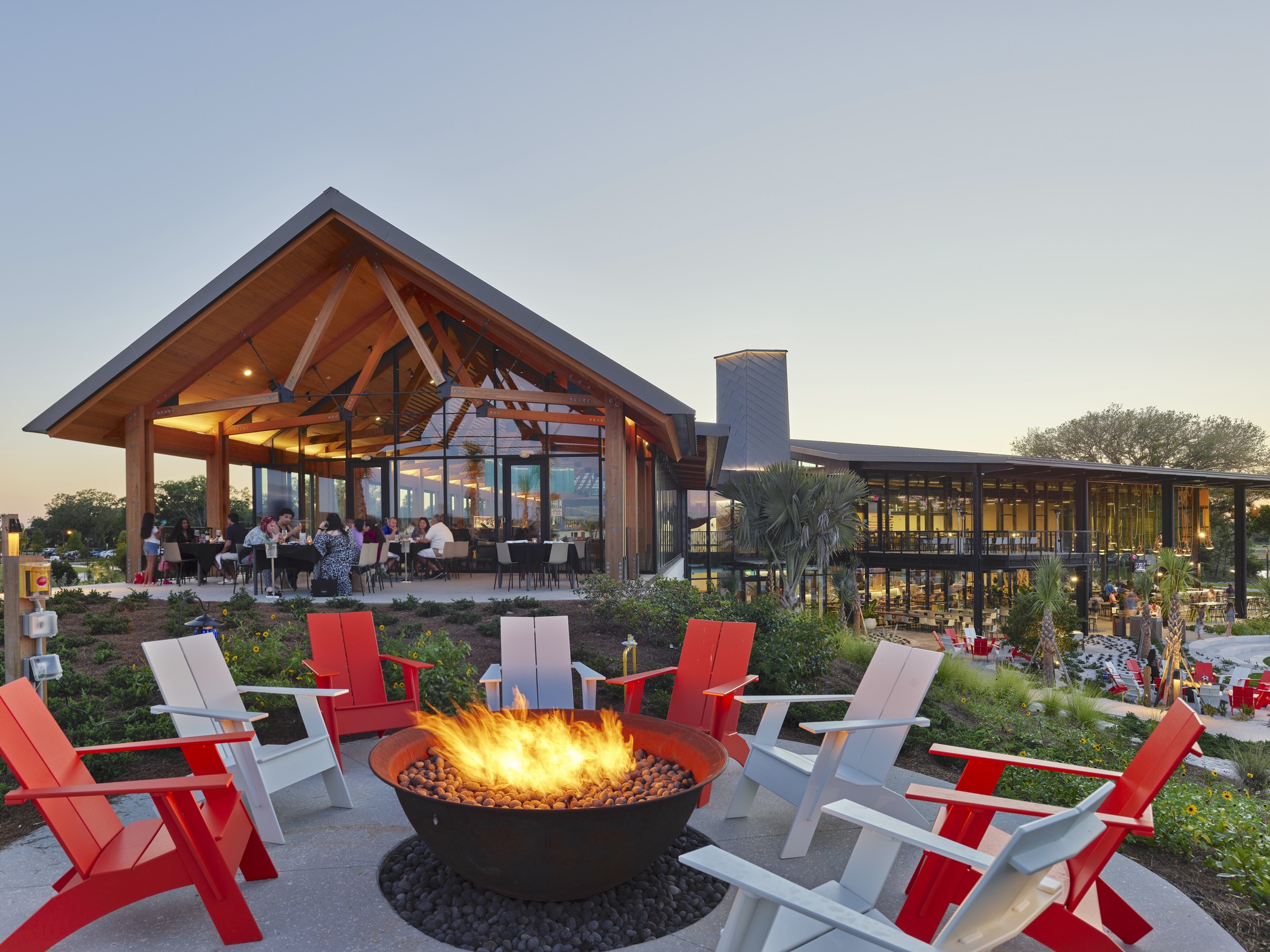 Outdoor fire pit surrounded by red and white chairs with covered dining pavilion at dusk