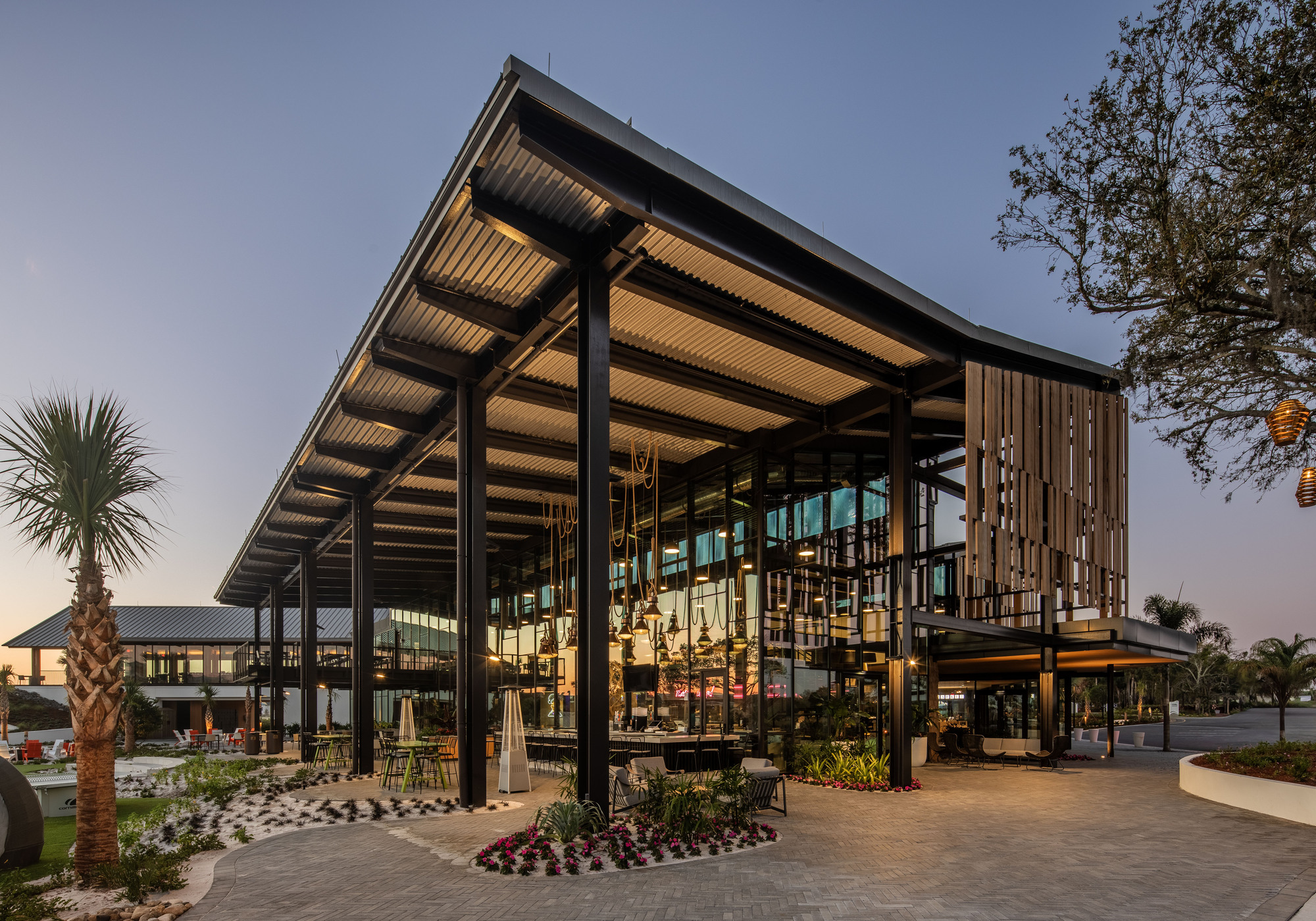 Covered pavilion with steel columns and slatted ceiling near planted palm trees at twilight