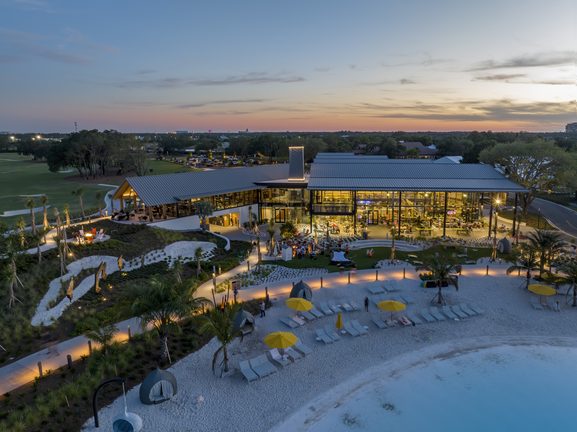 Aerial view of the poolside complex with sandy beach area and umbrella loungers at sunset