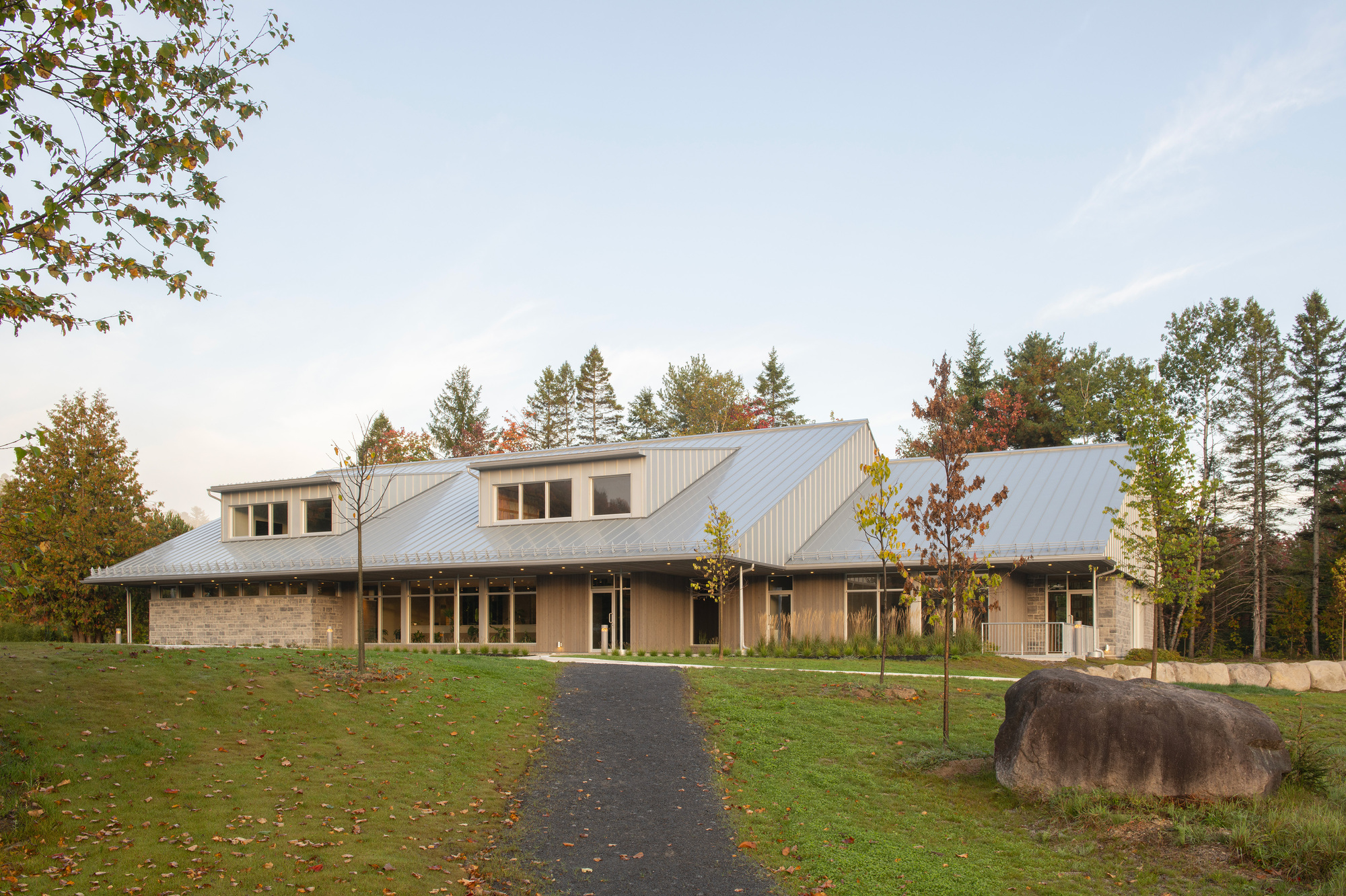Front elevation with stone masonry walls, dormers, and metal roofline approached by a paved drive