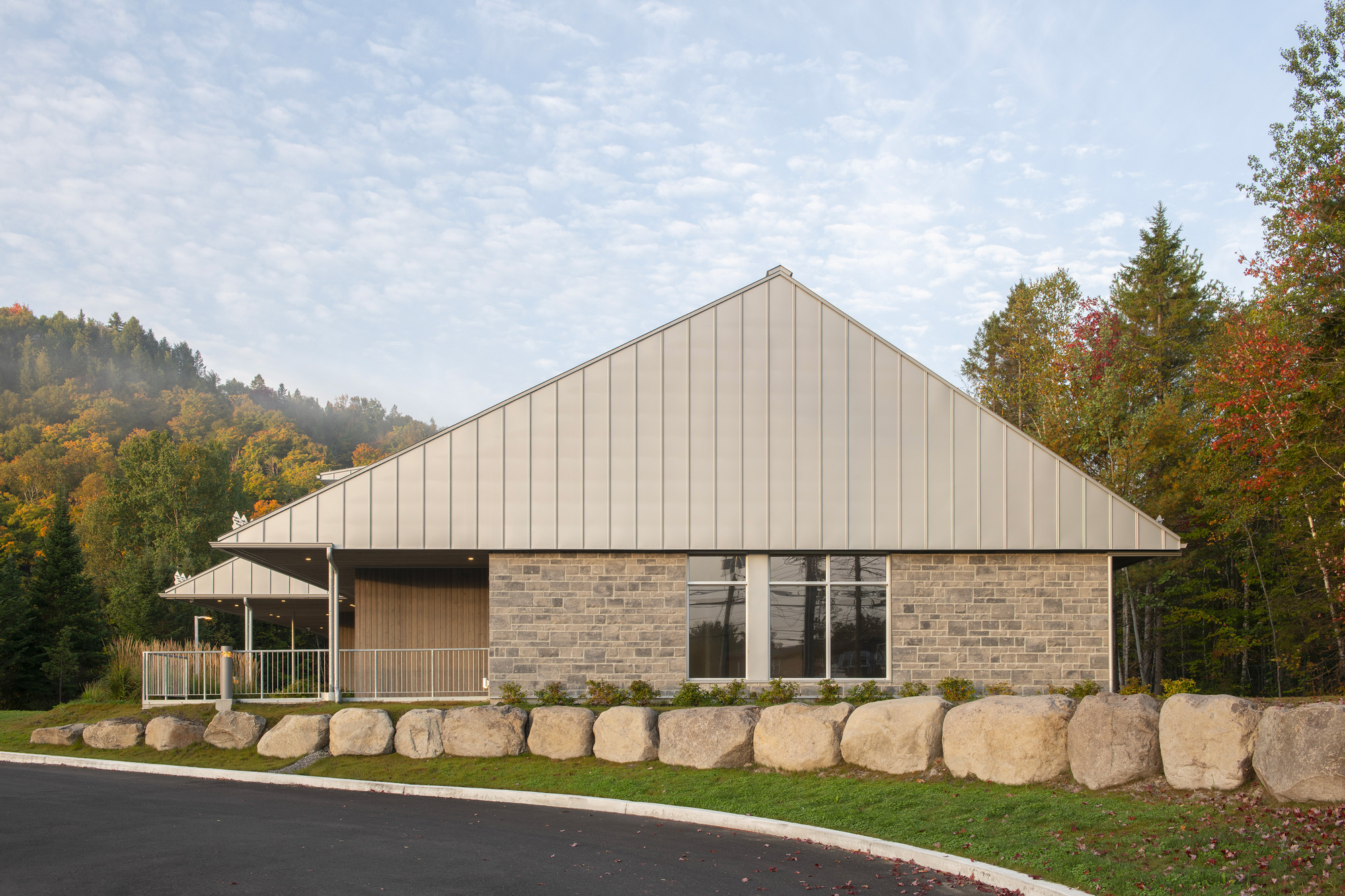Street facade with large boulder retaining wall, tall gable, and hillside forest beyond