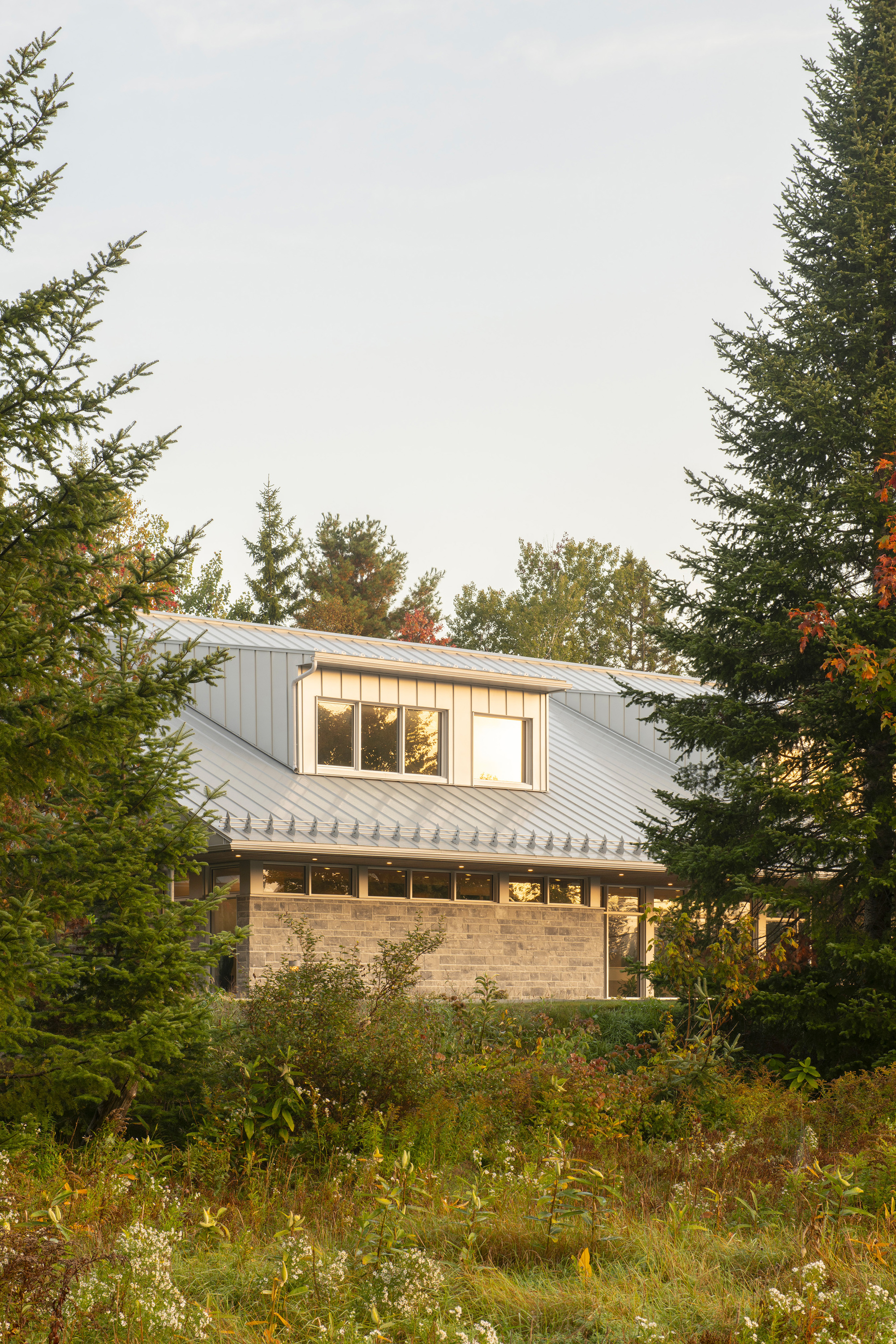 Rear view of the stone and vertical siding volumes framed by evergreen and deciduous trees
