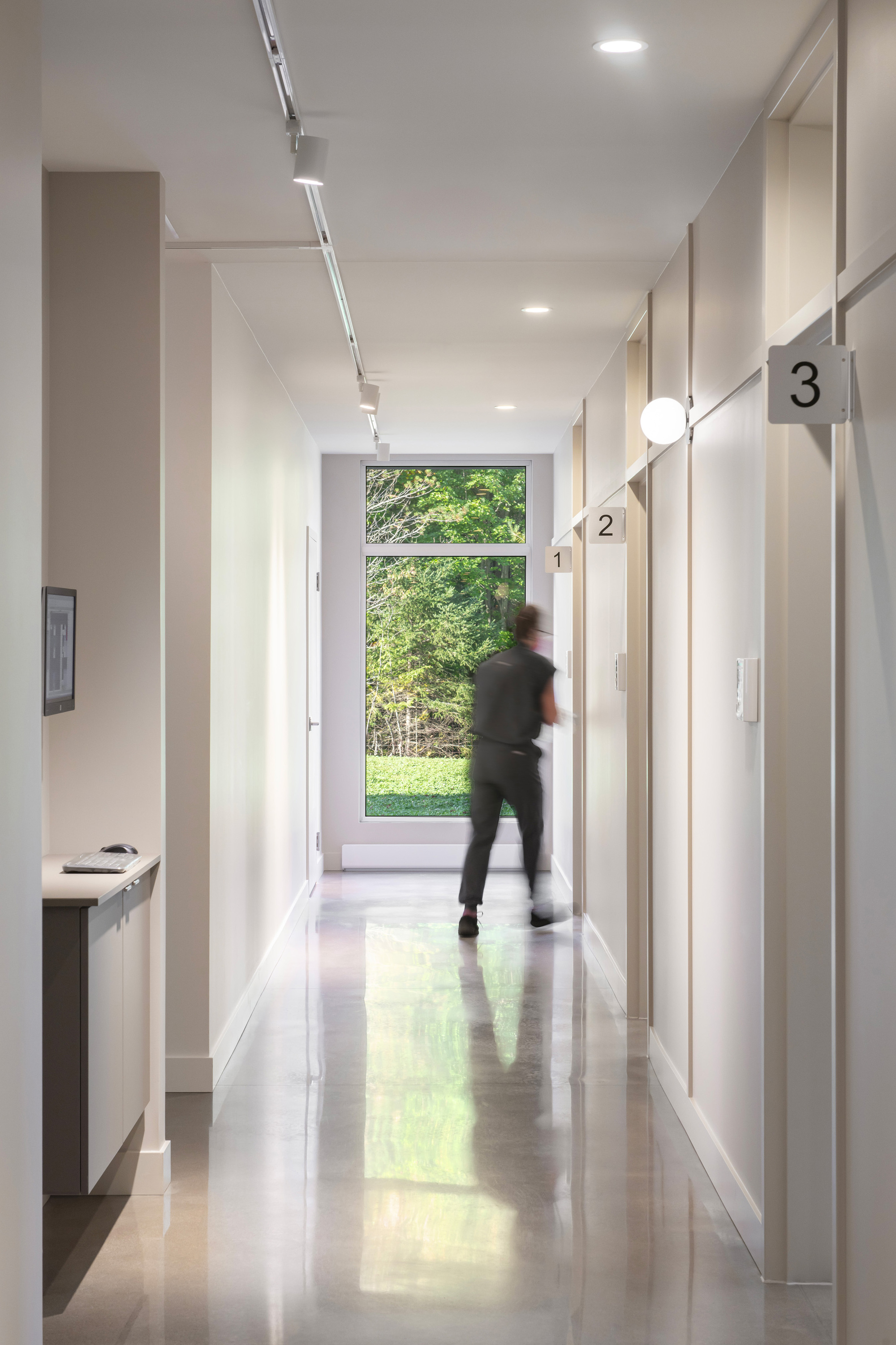 White corridor with polished concrete floor and figure in motion passing a large window to green trees