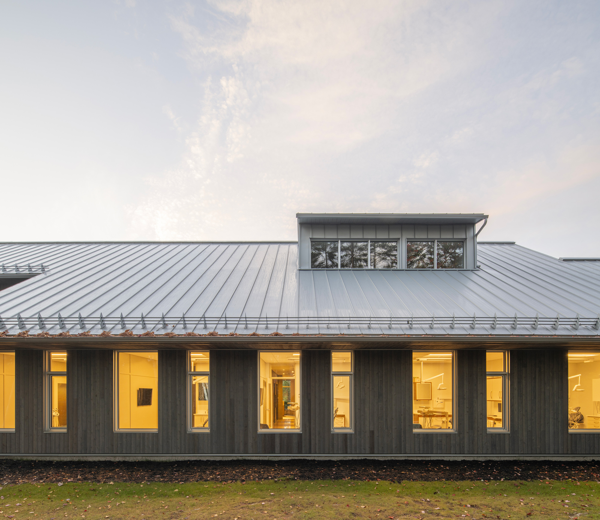 Dark wood facade with vertical window openings and clerestory dormer beneath standing seam metal roof at dusk