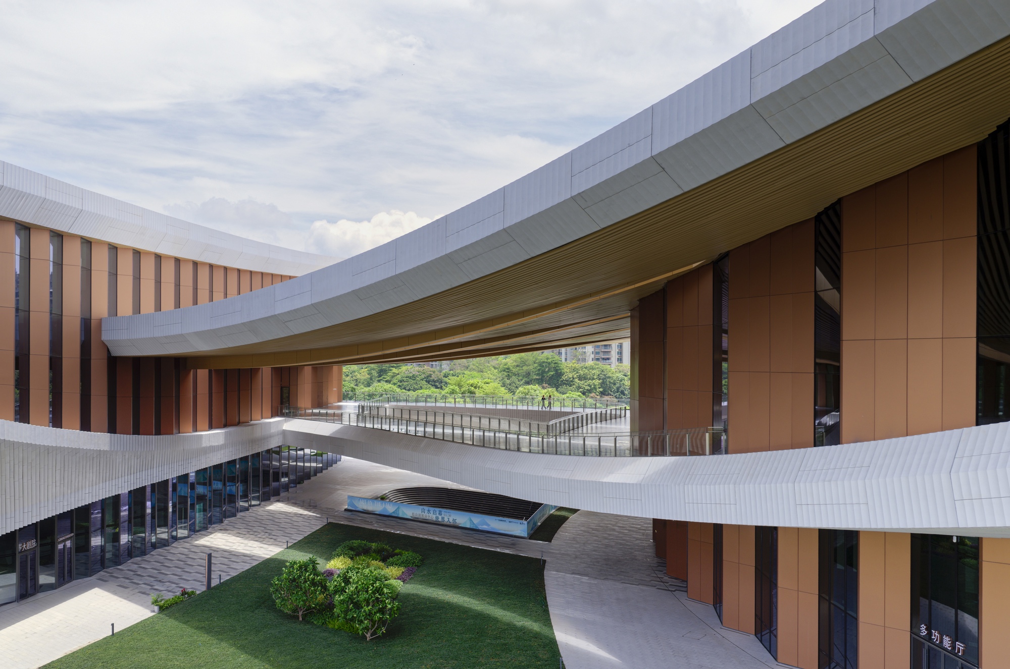 Courtyard view framing the white pedestrian bridge spanning between timber-clad walls and vertical louvers