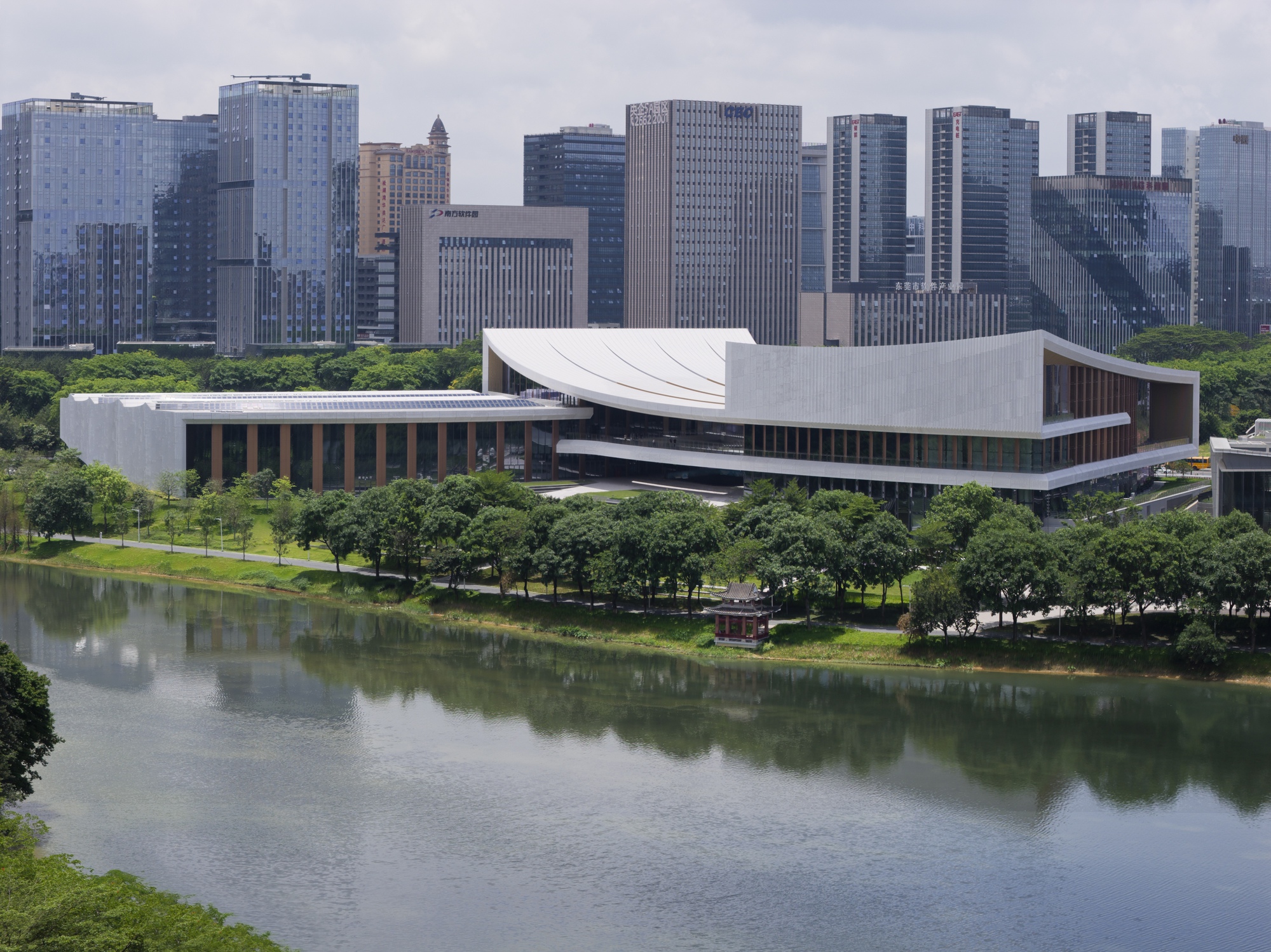 Elevated view of the white volume with curved roof beside a pond and park landscape with high-rise towers beyond