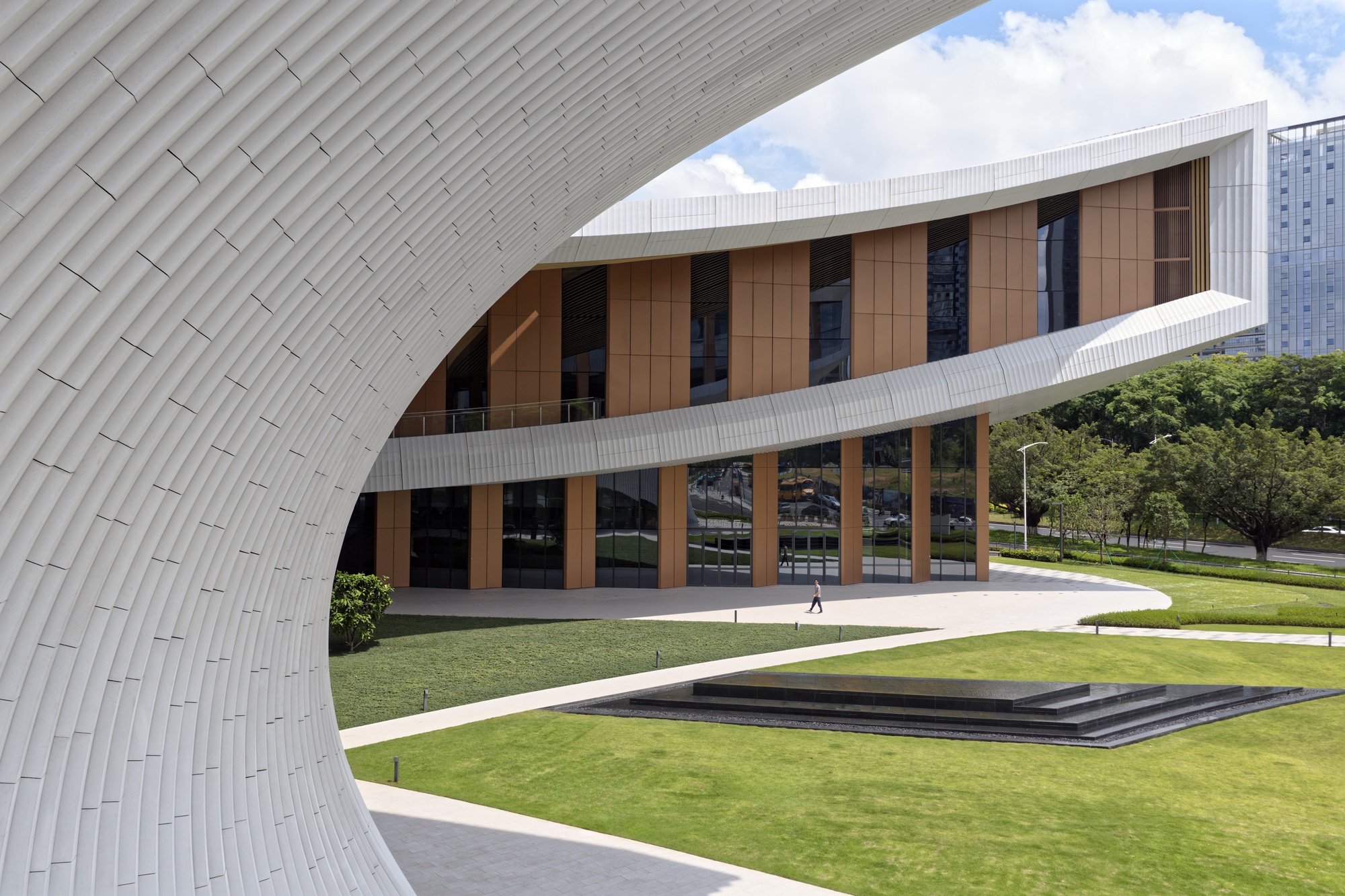 Curved white tile wall wrapping past glazed facade and stepped water feature in manicured grass courtyard
