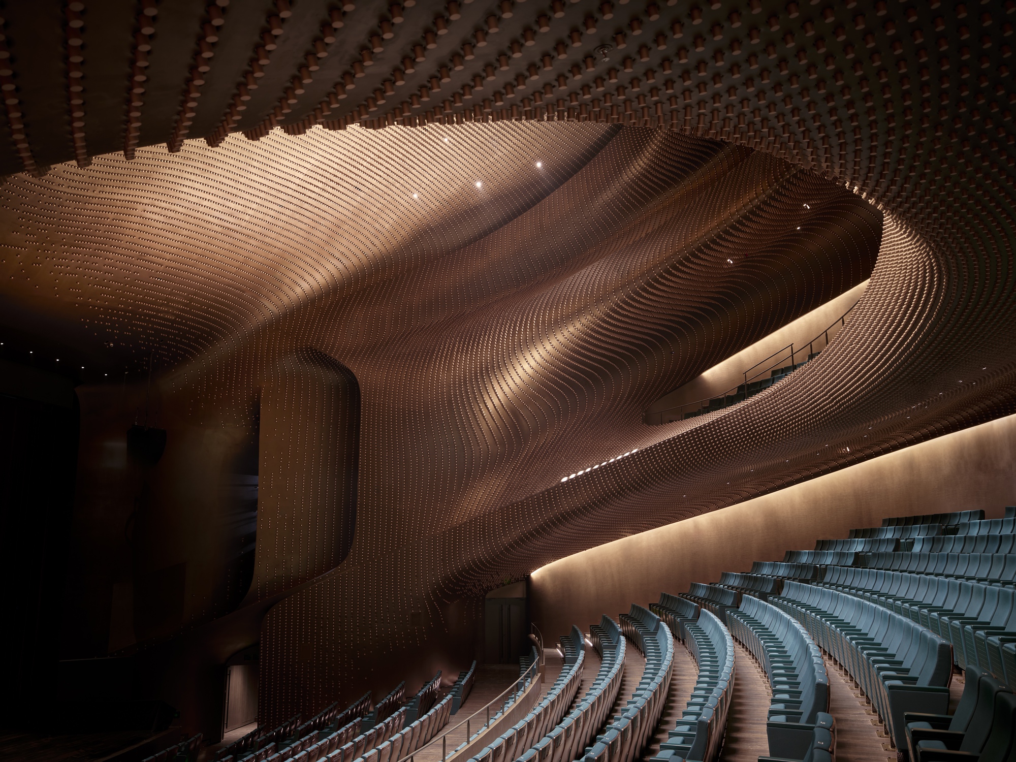 Auditorium interior with undulating timber ceiling and rows of blue seats under theatrical lighting