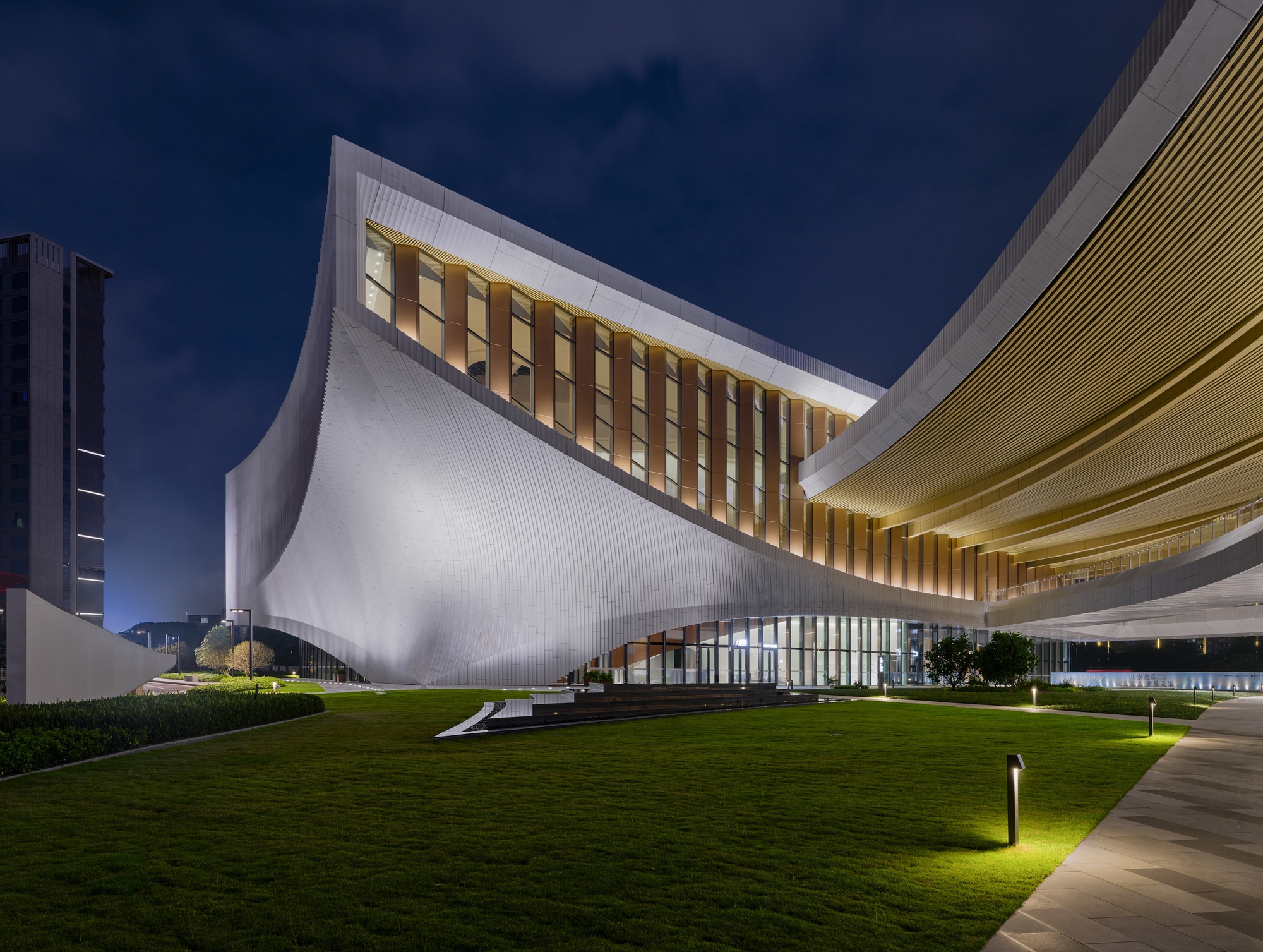 Curved white facade with vertical louvers rising above a grassy lawn at twilight