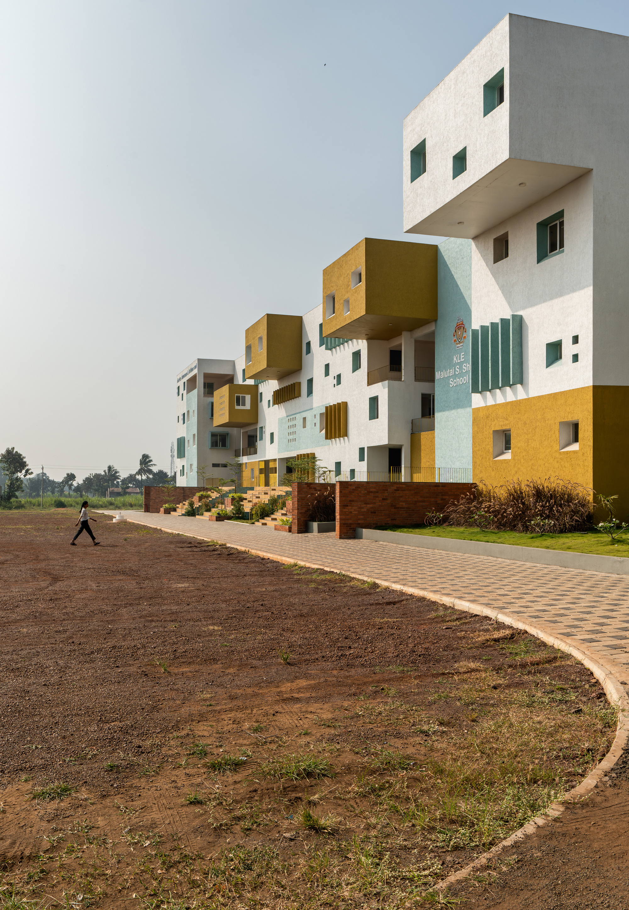 Stacked volumes clad in white, yellow and pale blue with cantilevered boxes and a person crossing the brick paved forecourt