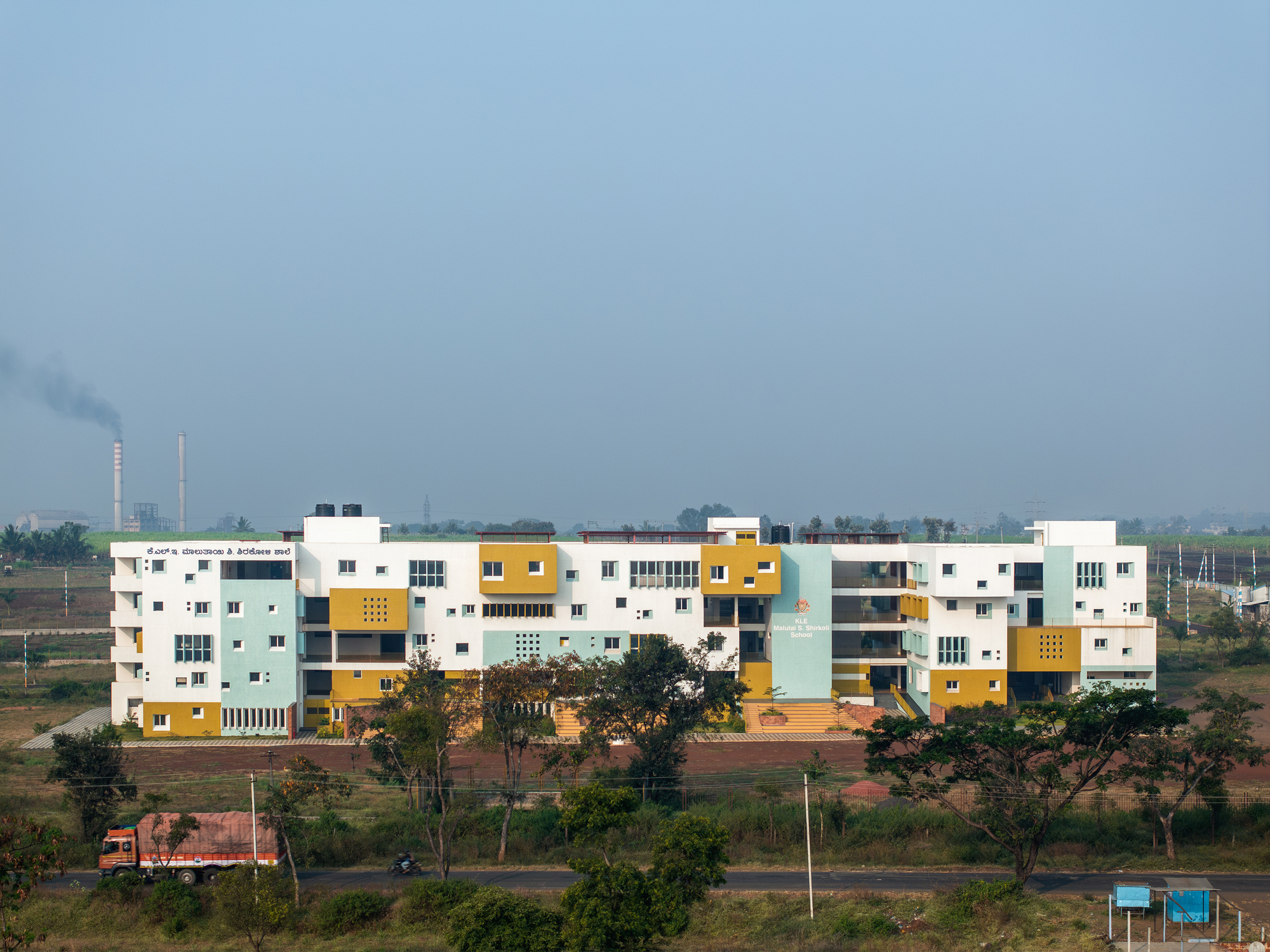 Horizontal facade of interlocking volumes in white, yellow and pale blue under a hazy sky with distant smokestacks