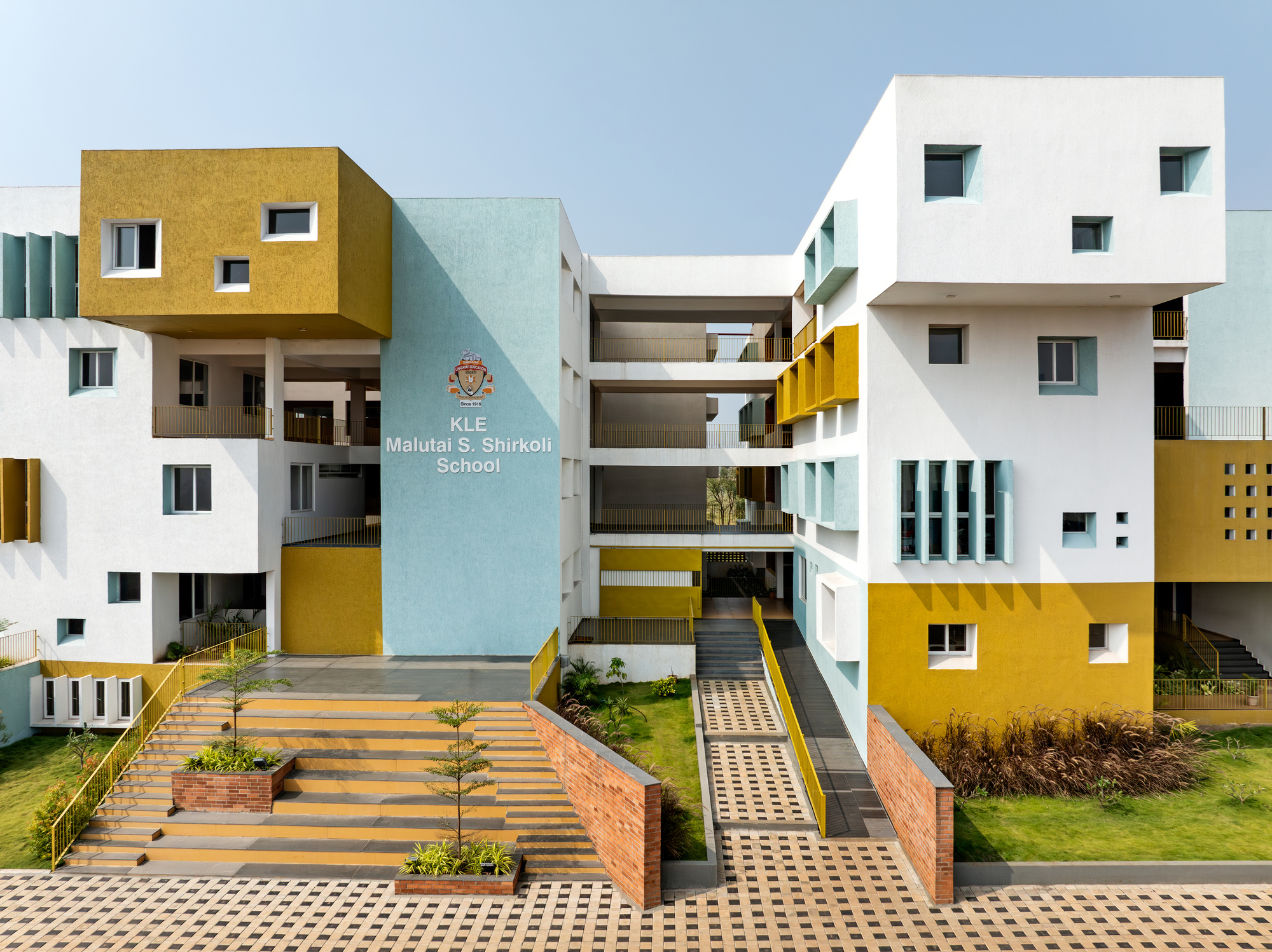Entrance court with wide brick steps, planted beds and tiered facade volumes in yellow, white and pale blue