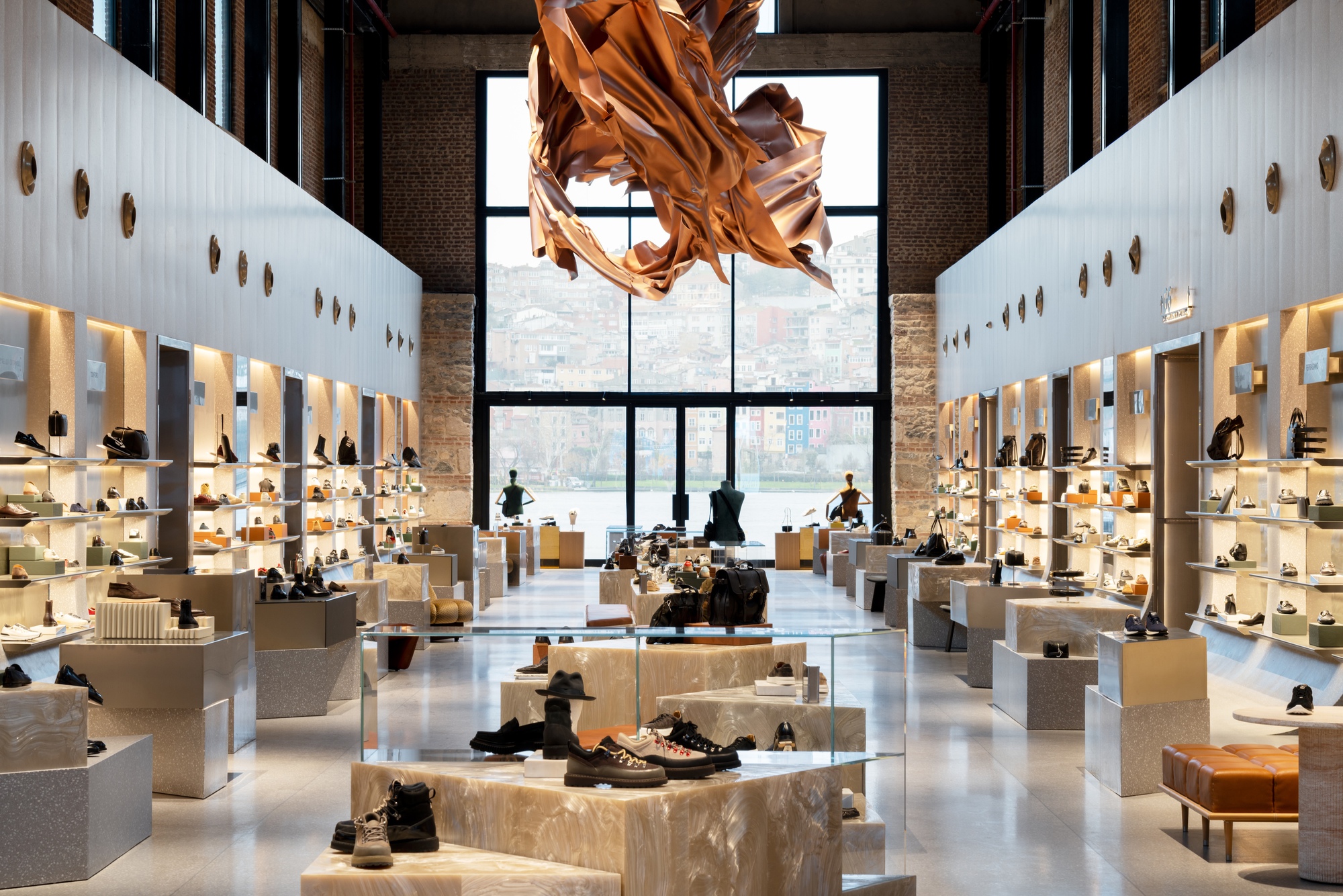 Double height retail hall with white shelving walls and copper fabric sculpture suspended before the glazed facade