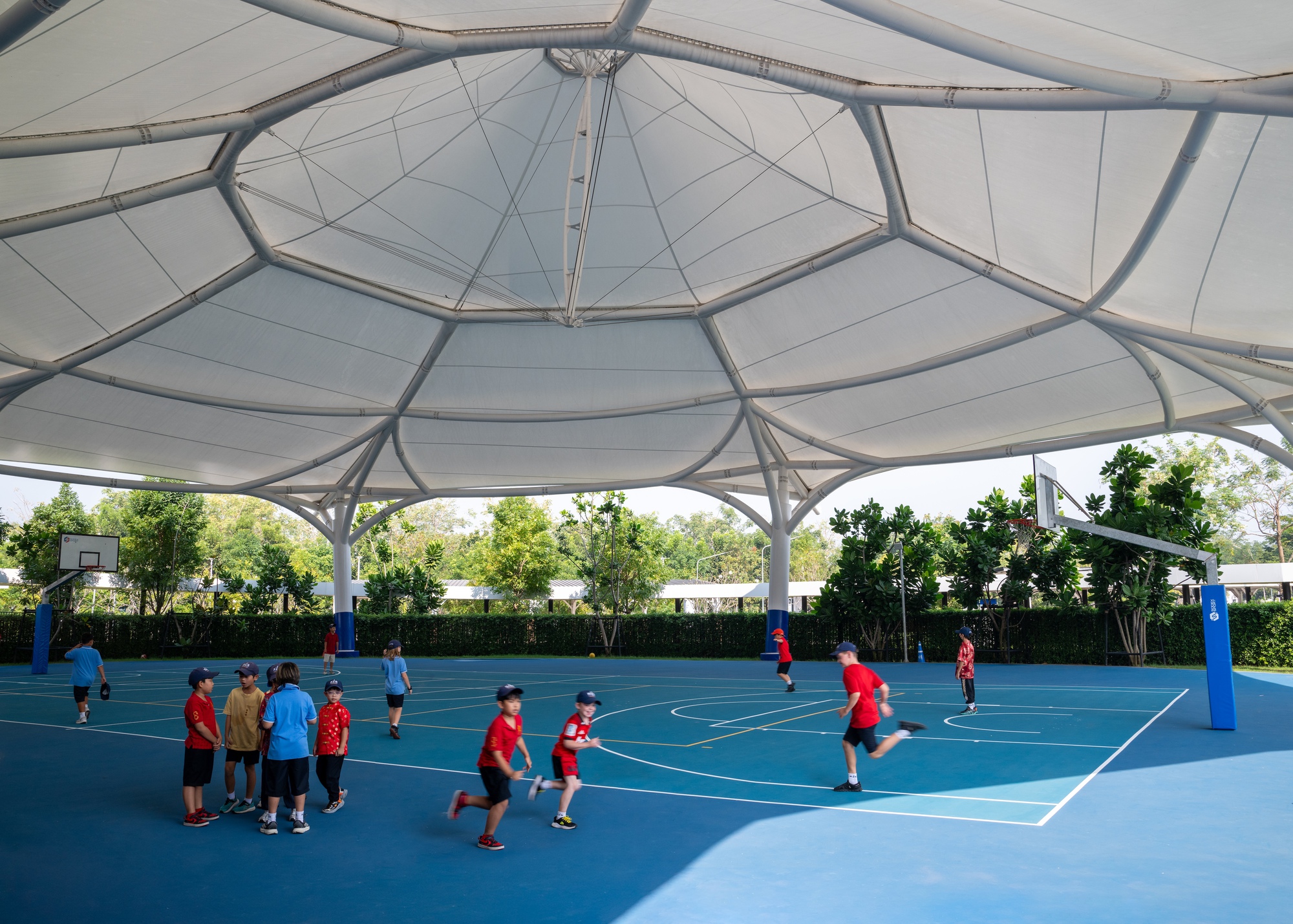 Fabric tensile canopy sheltering a basketball court where students play beneath steel support rings