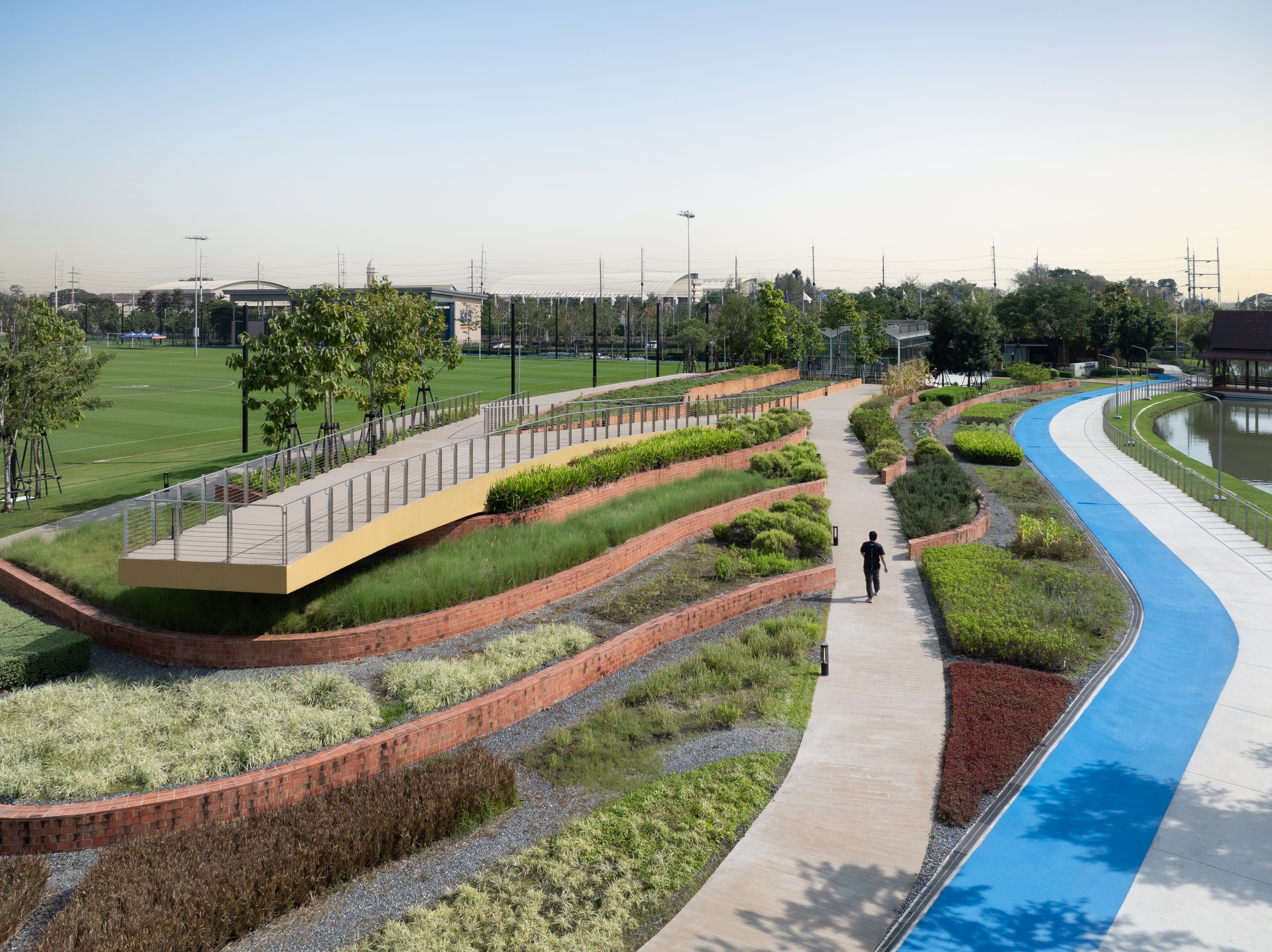 Curving pathways with blue running track and planted beds stretching past sports fields at dusk