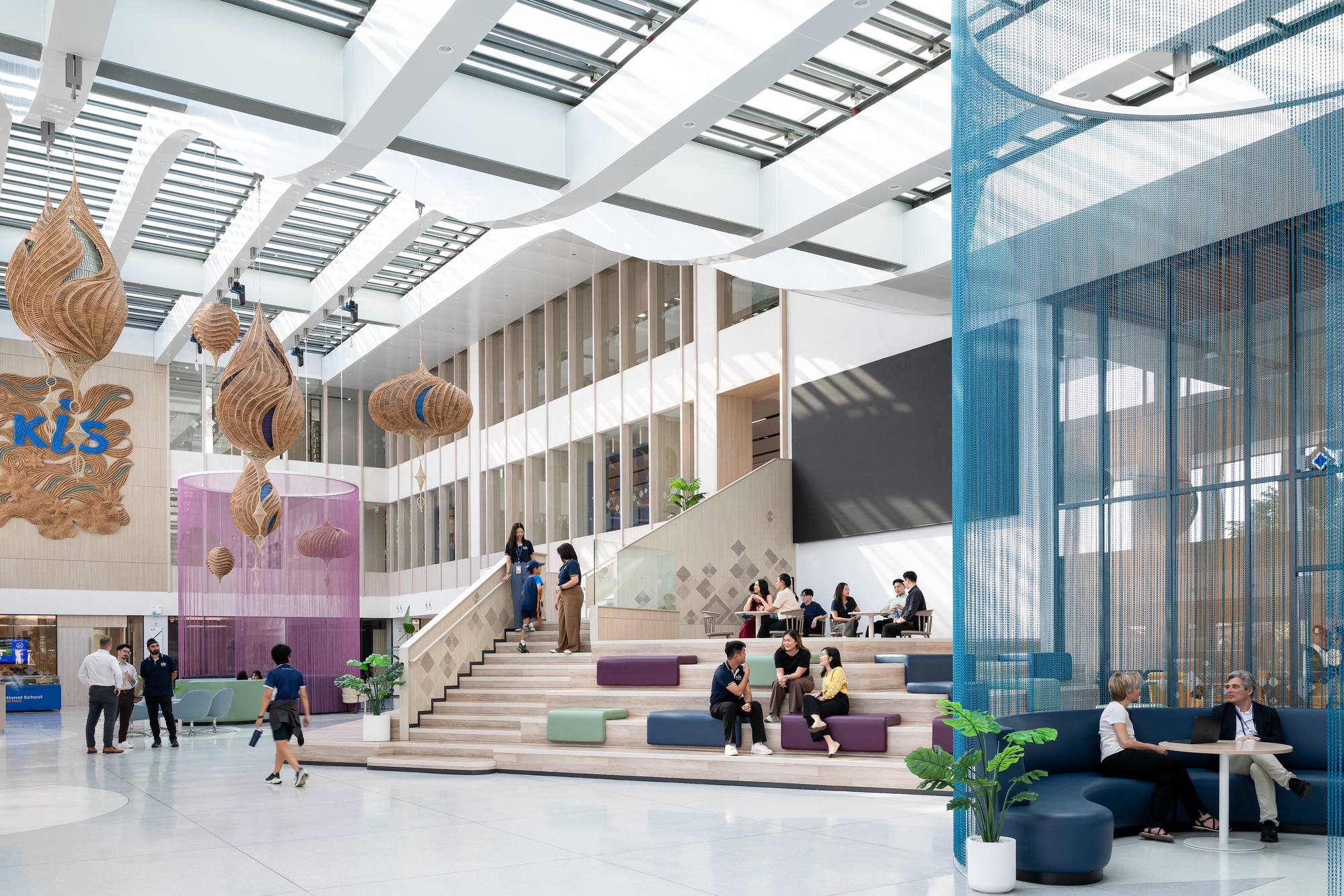 Double-height atrium with tiered seating steps, woven pendant lights, and students gathered throughout the space