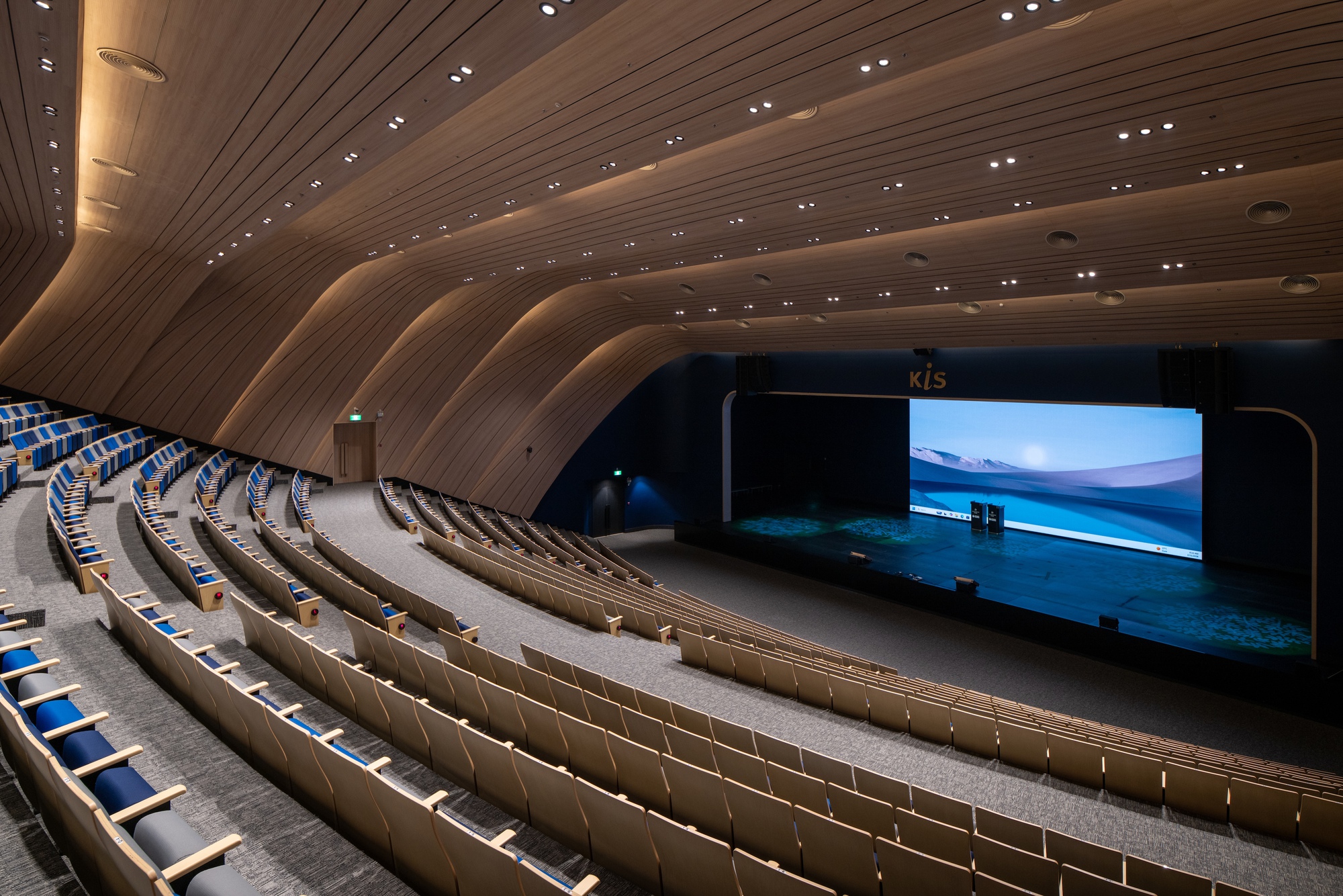 Auditorium interior with curved timber-clad ceiling and tiered seating facing an illuminated stage screen