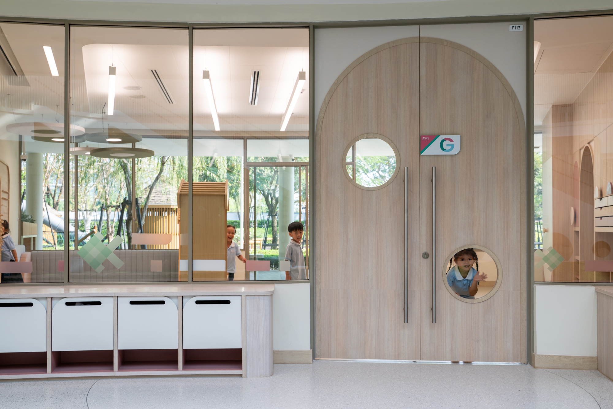 Arched timber door with circular portholes looking into a glass-partitioned corridor with children passing through