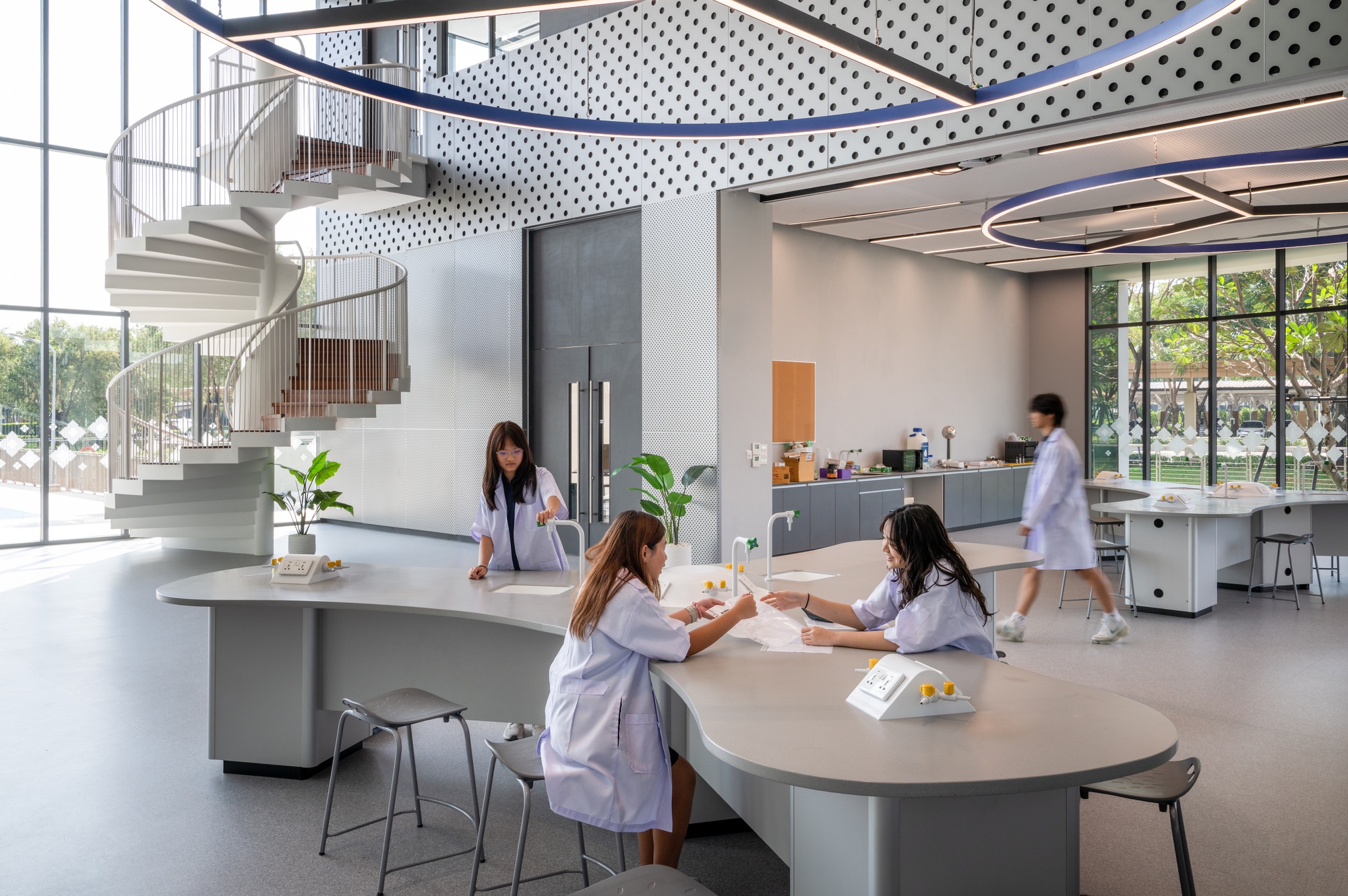 Open workspace with curved tables and spiral staircase beneath perforated ceiling panels with students in white coats