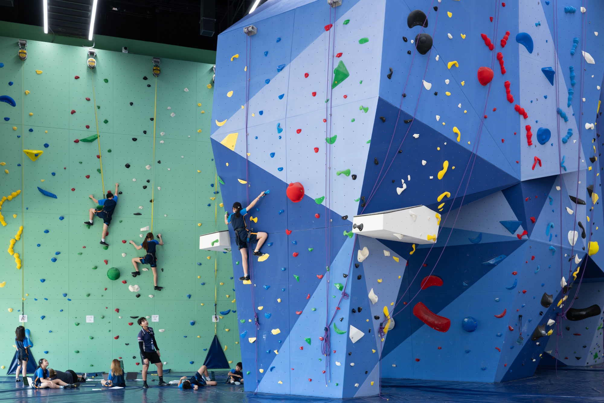Indoor climbing wall with multicolored holds on angled blue and green panels with climbers scaling the surface