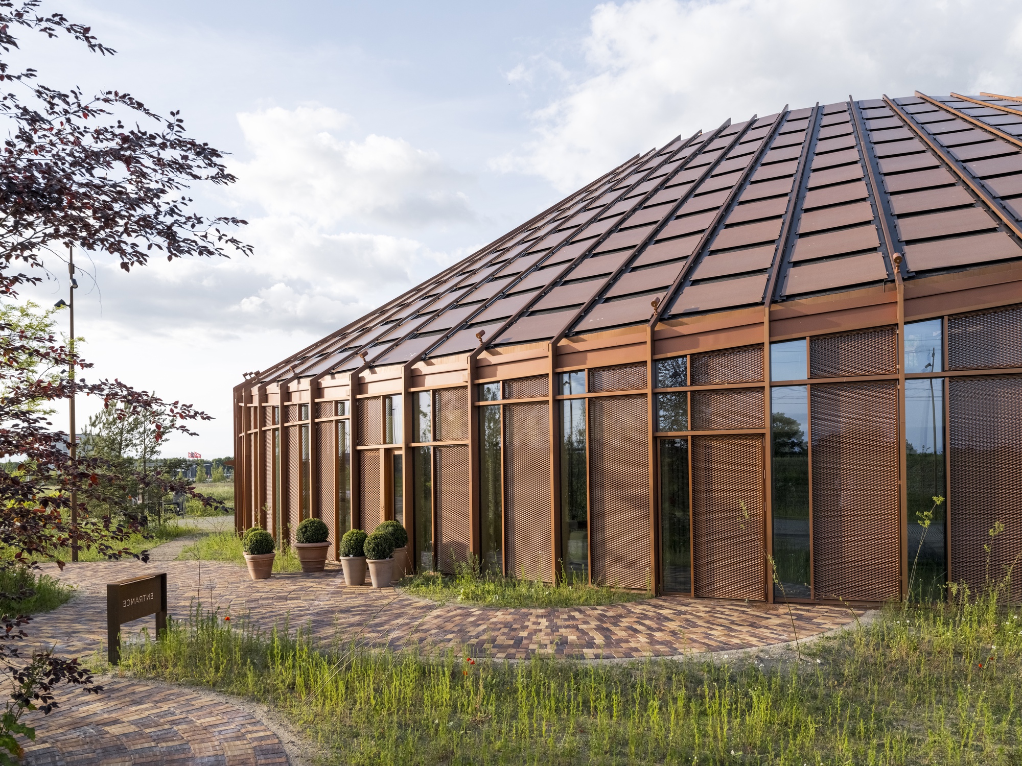 Entrance facade with copper panel roof and vertical timber-framed glazing beside a brick pathway and potted shrubs
