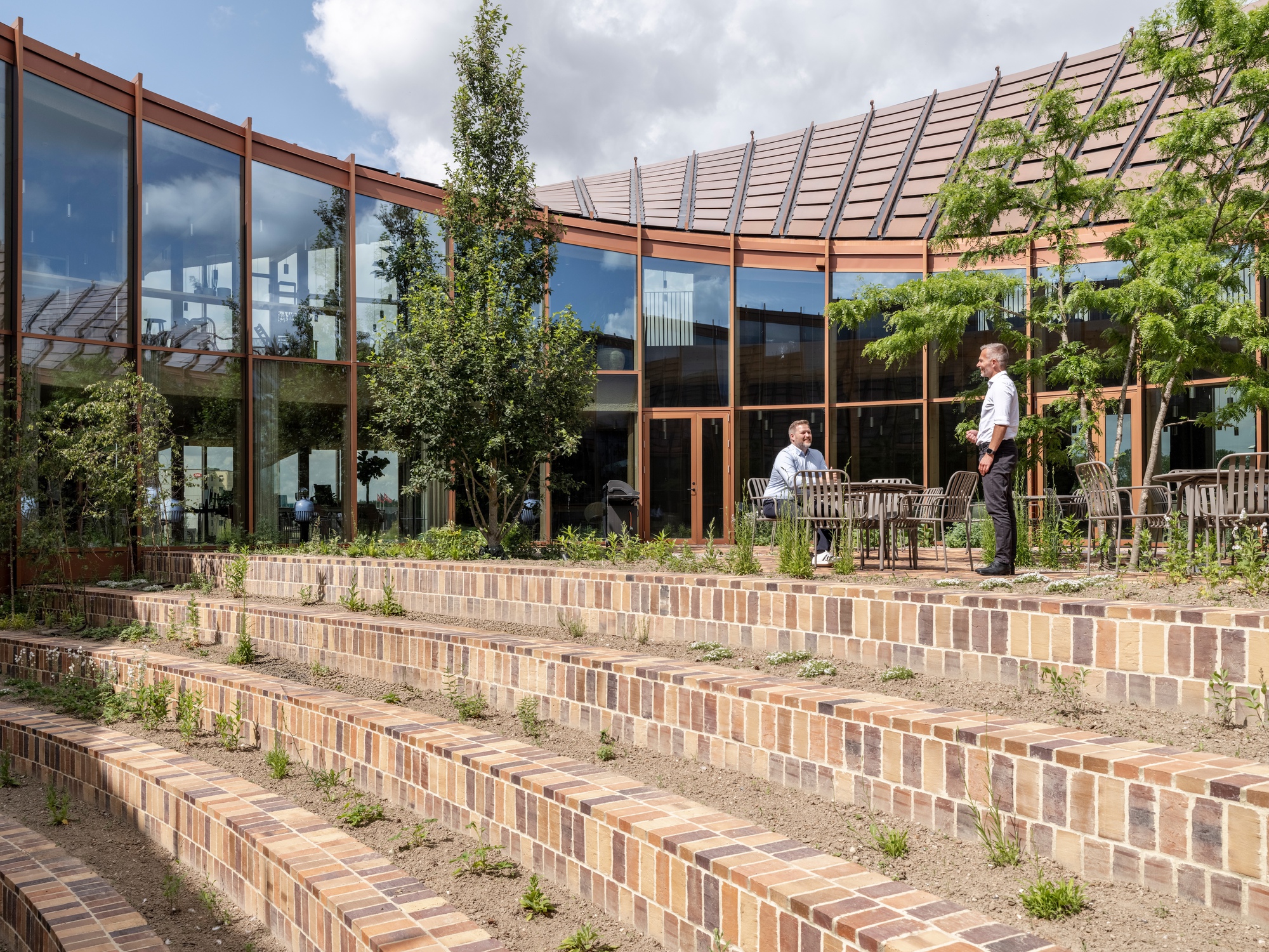 Tiered brick seating steps with young trees in front of a glazed facade with timber frame and sawtooth roof