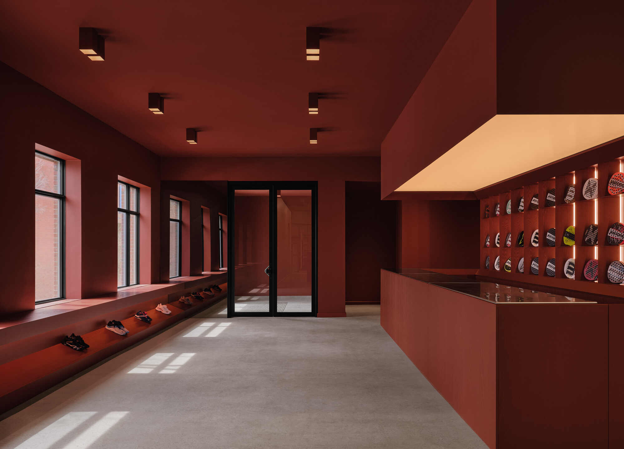 Gallery space with deep red walls, recessed shelving, and tall windows casting afternoon light across polished concrete