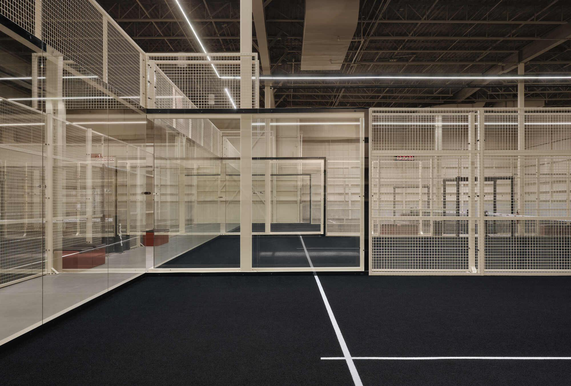 Indoor sports court enclosed by white metal mesh partitions beneath an exposed joist ceiling with linear lighting