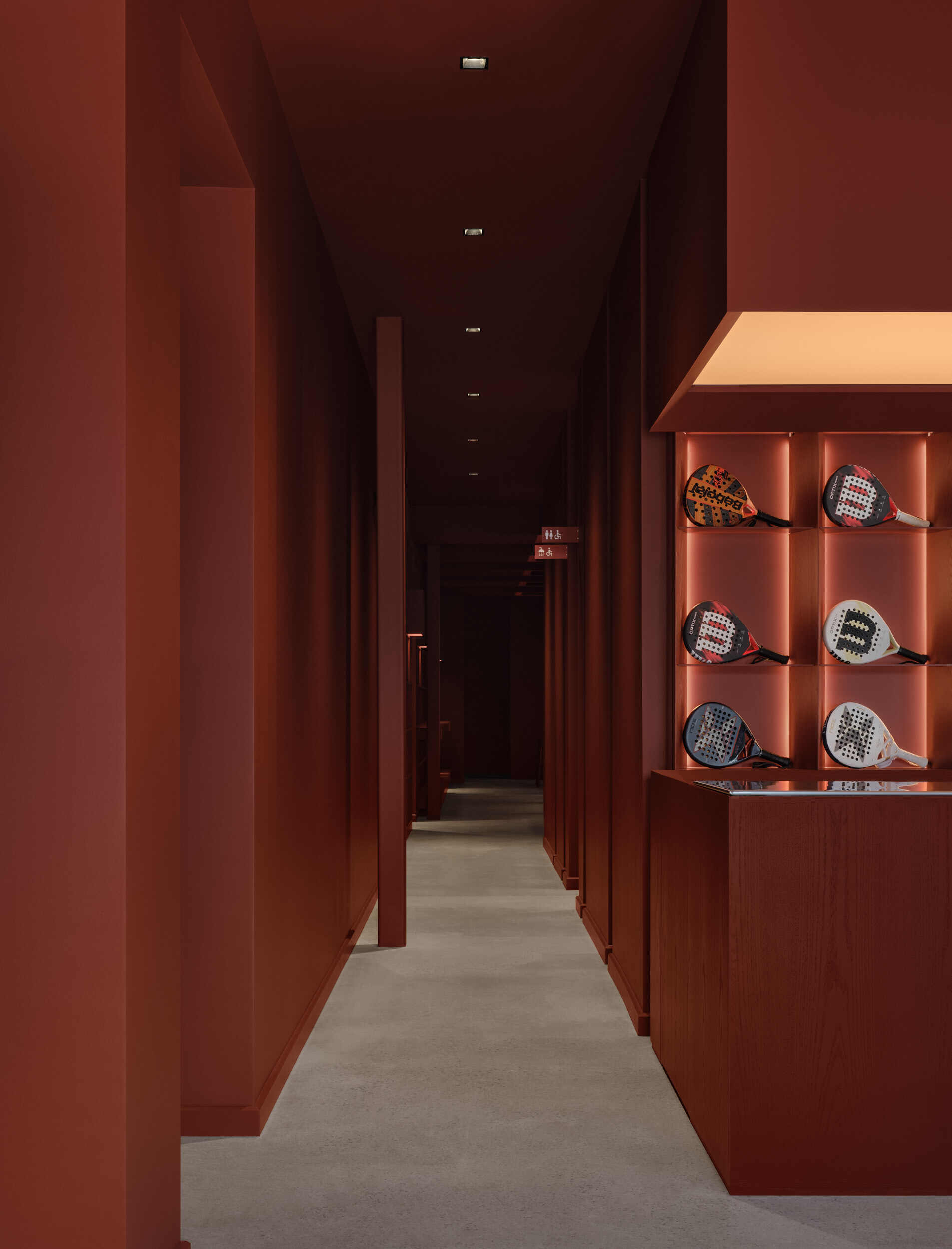 Narrow corridor with terracotta-paneled walls, concrete flooring, and display niches containing sports equipment