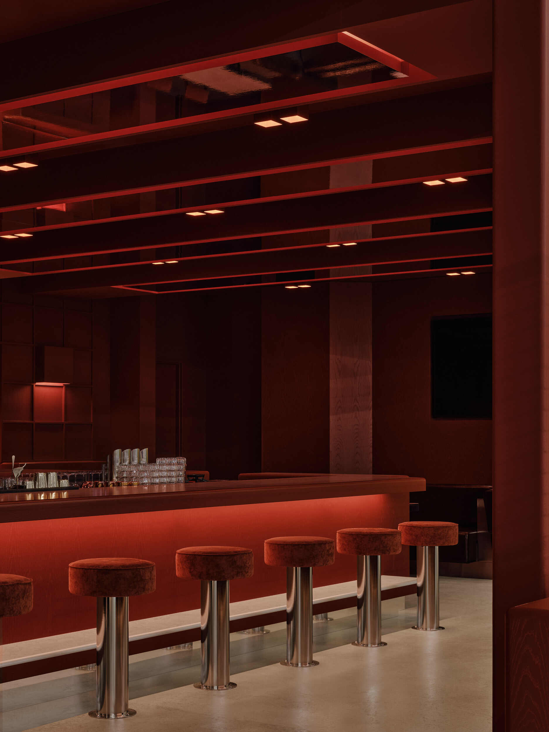 Bar counter with upholstered stools on metal pedestals beneath layered terracotta ceiling beams and recessed lighting