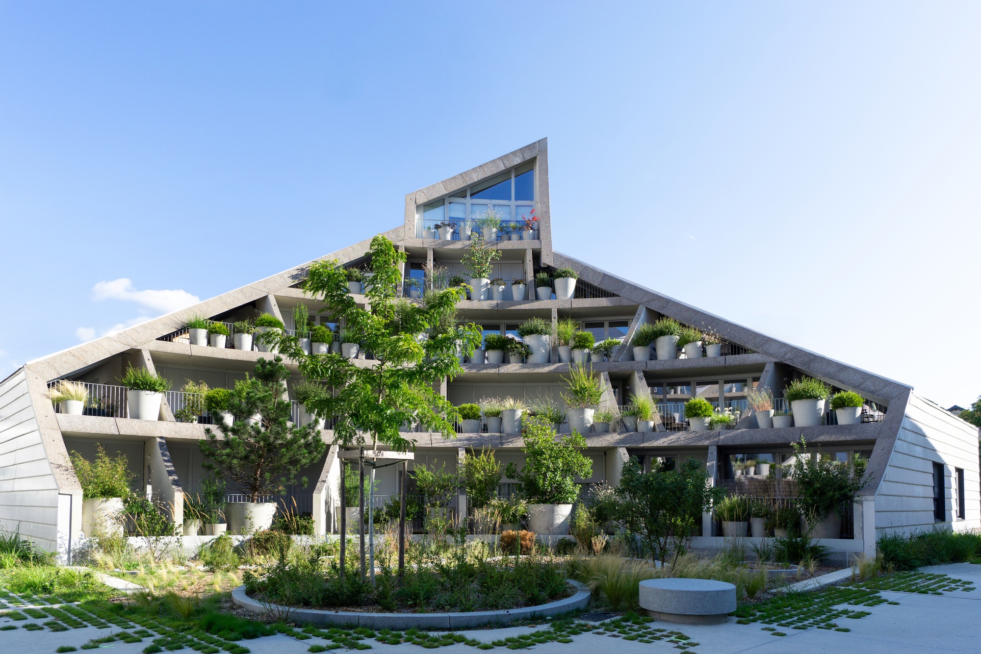 Angled facade with planted terraces surrounding a paved courtyard with grid pattern and small trees