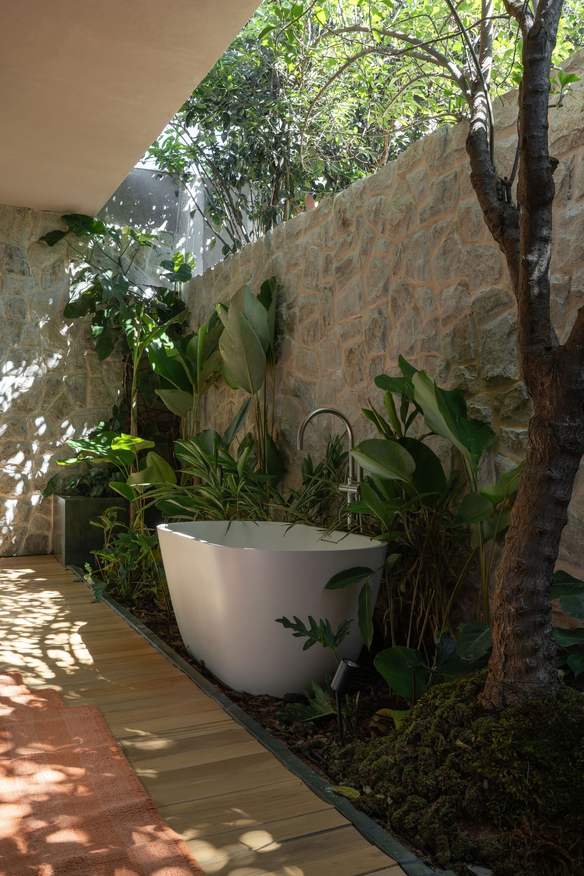 Freestanding bathtub in open-air courtyard with stone walls and tree canopy overhead
