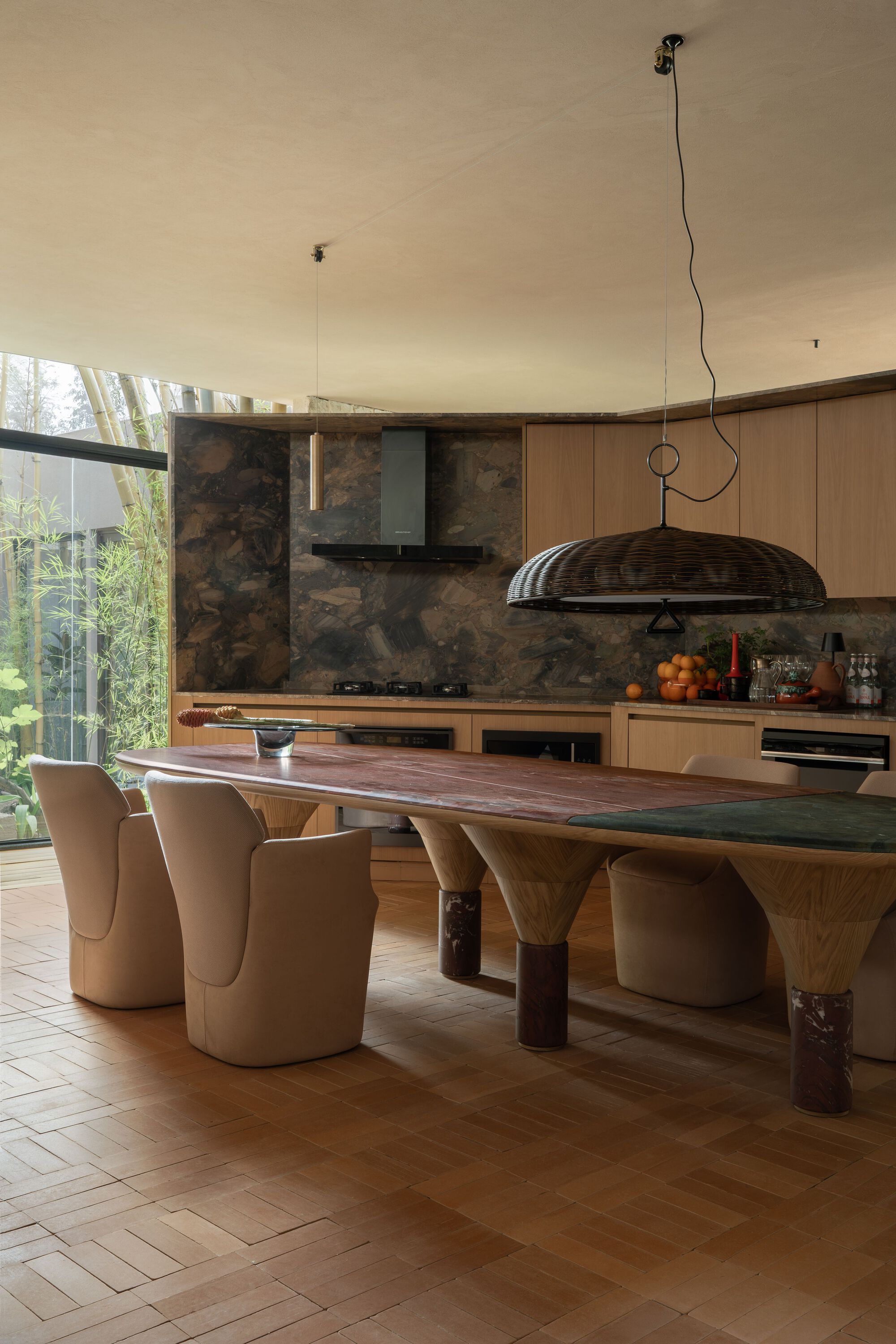 Sculptural dining table with curved timber legs below a woven dome pendant near glazed garden views