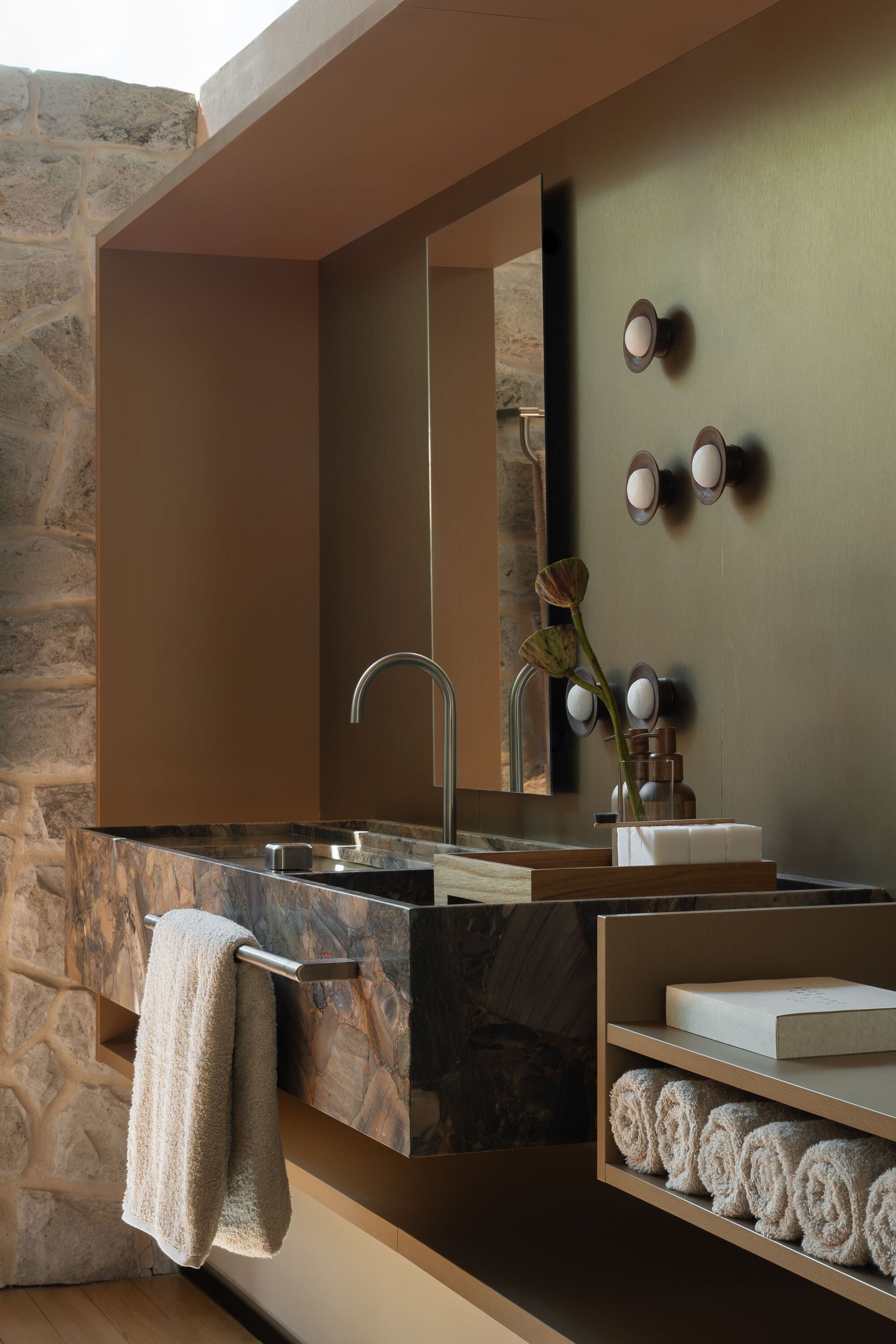 Bathroom vanity with dark marble counter, mirror and timber storage against stone wall with circular sconces