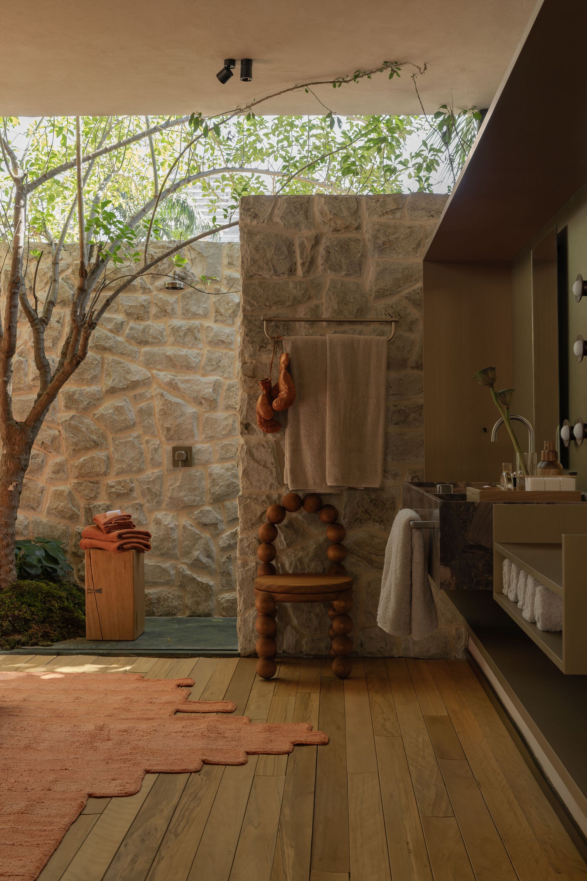 Open-air bathroom with stone walls, timber bathtub and tree branches overhead filtering daylight