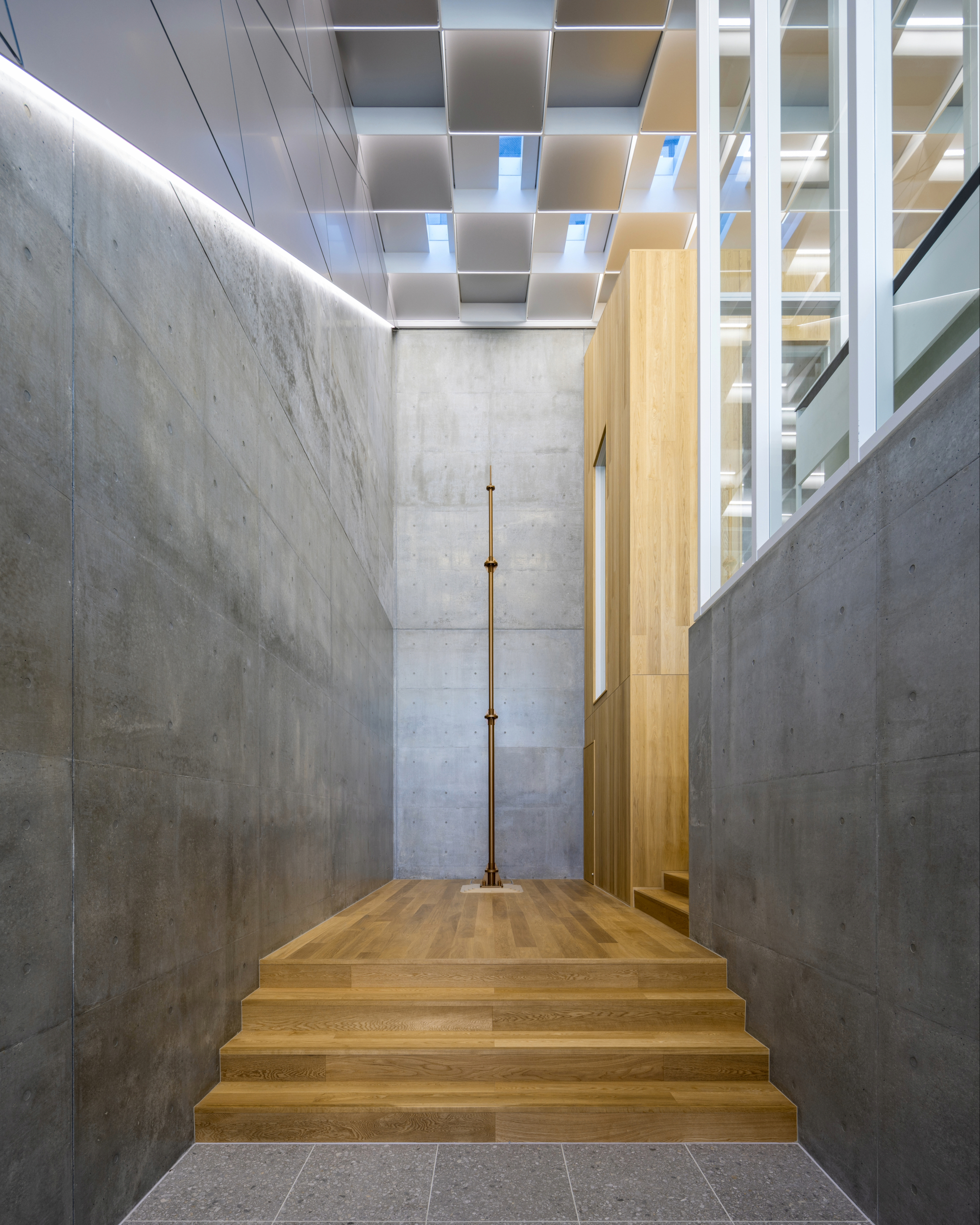 Concrete walled corridor with timber stairs rising toward geometric coffered ceiling and natural light