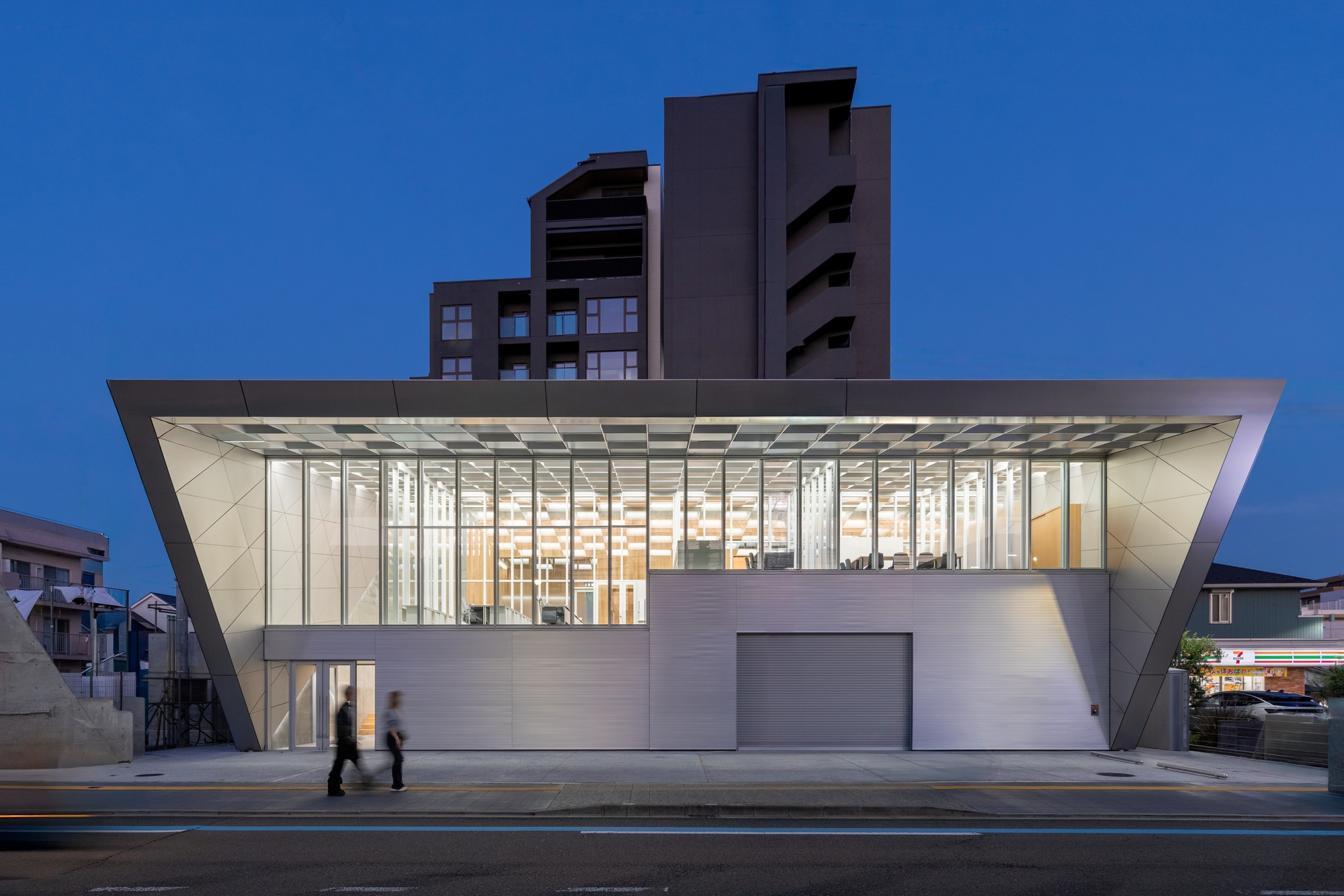Street facade with cantilevered glass volume above horizontal cladding at dusk with pedestrians passing