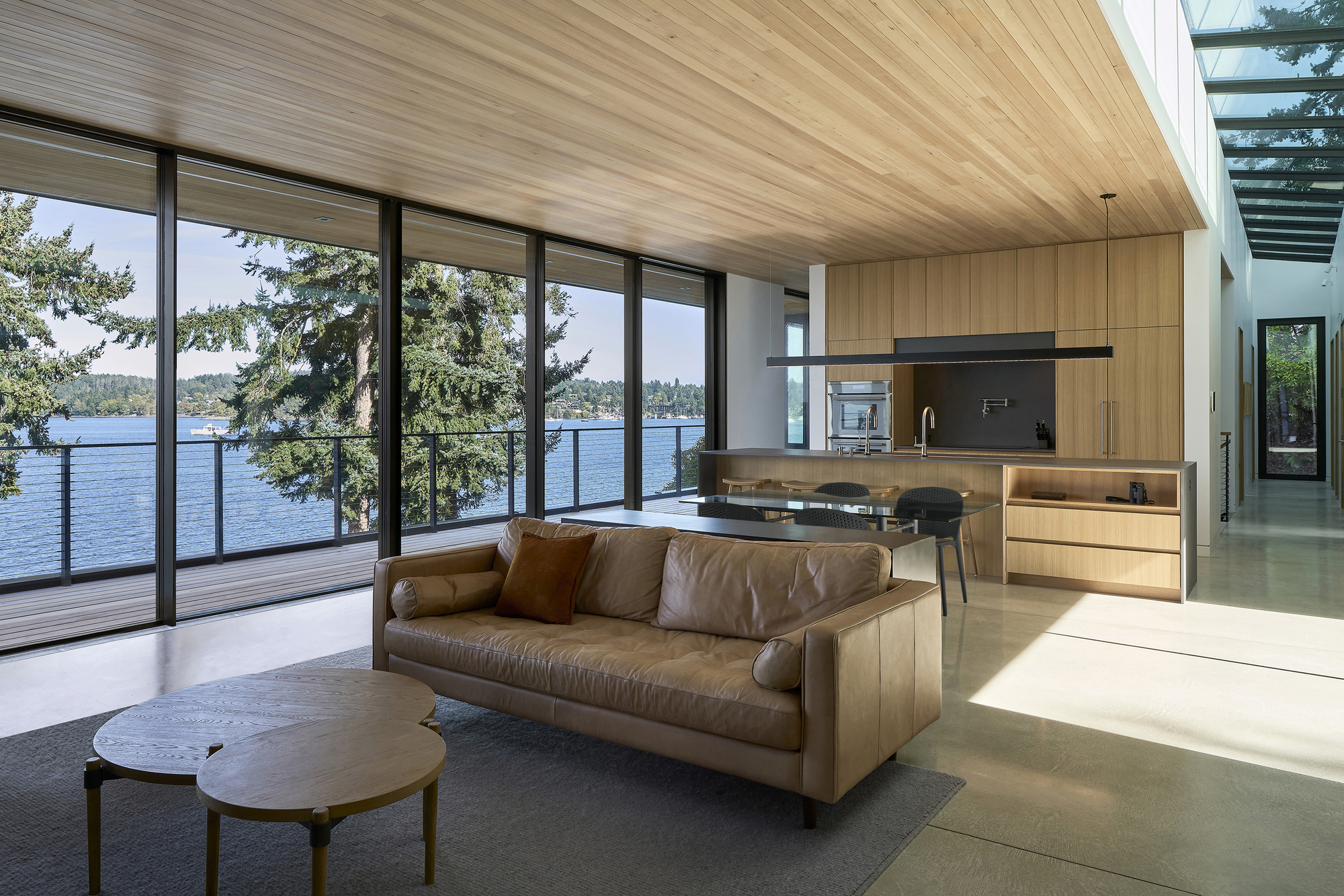 Open living space with timber ceiling and floor-to-ceiling glazing overlooking a lake through evergreen trees