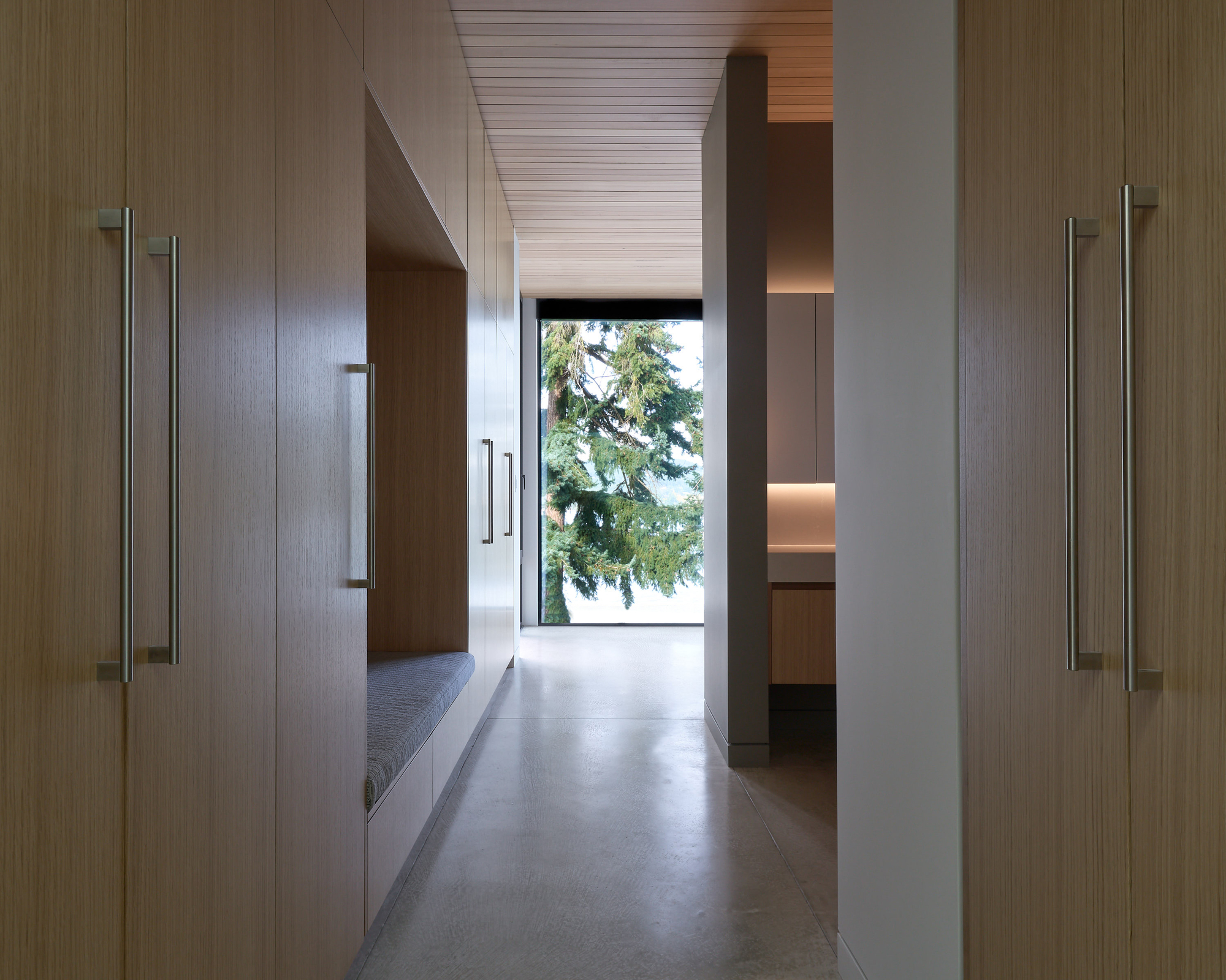 Narrow hallway lined with wood-paneled walls and concrete flooring leading to a framed view of evergreen trees