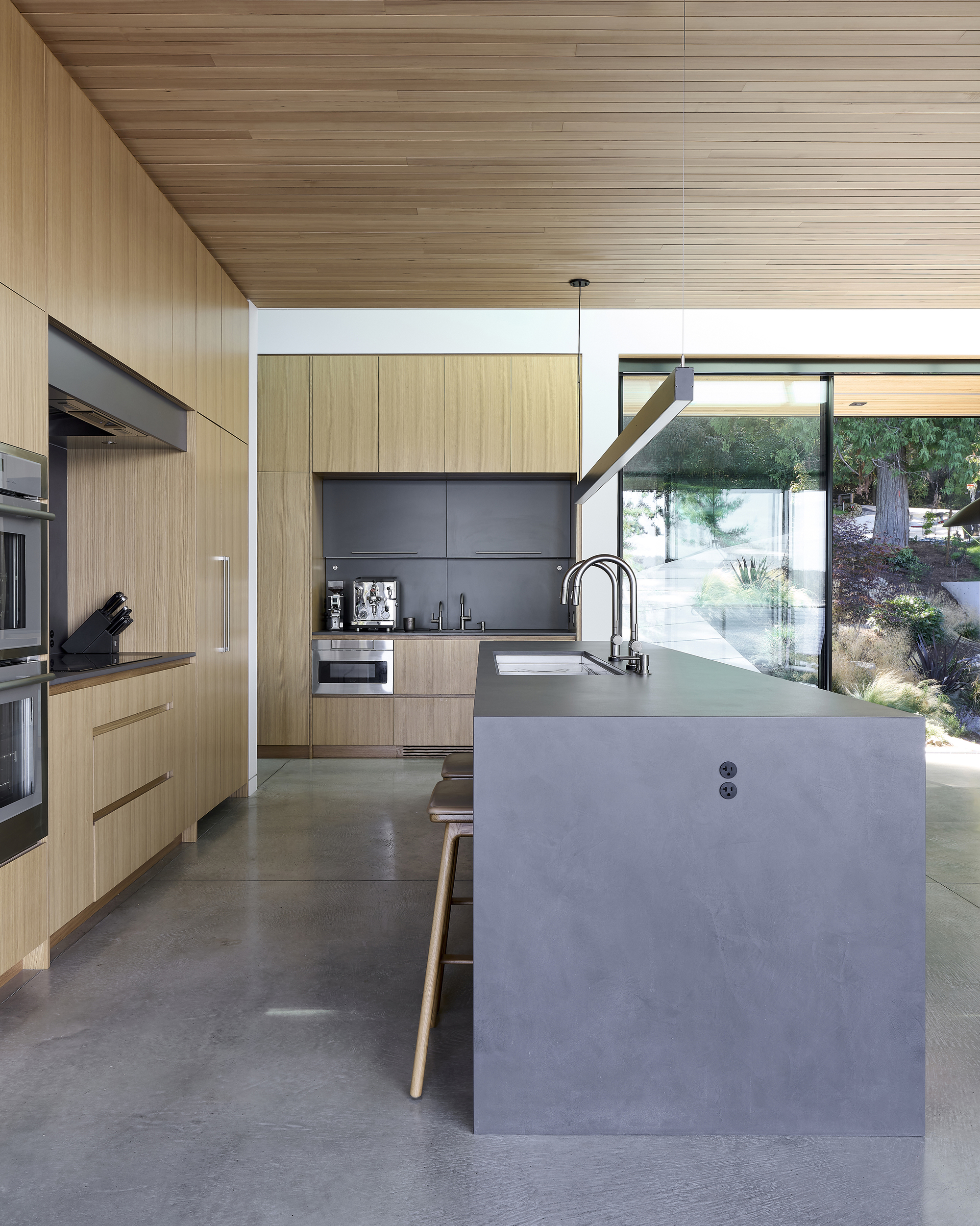 Kitchen island with pendant light fixture beneath wood-paneled ceiling overlooking a sloped garden through glazed walls