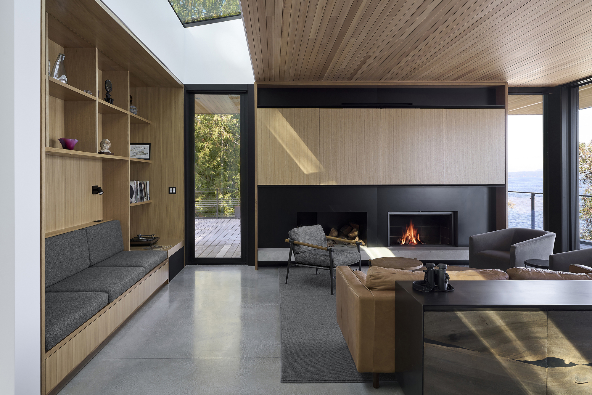 Living room with timber shelving wall, steel fireplace, and skylights casting afternoon light on concrete floors