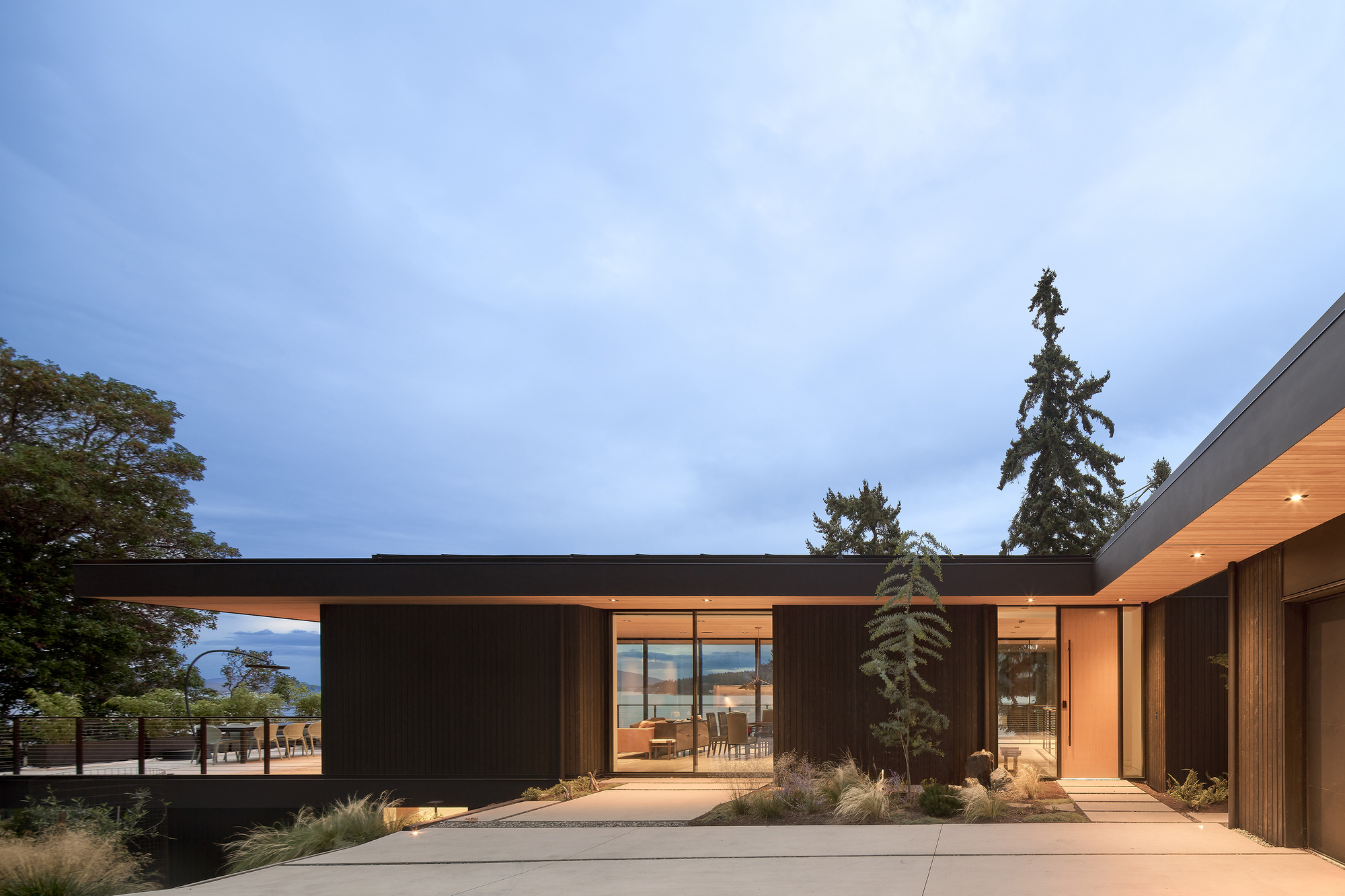 Courtyard entry with dark vertical cladding, timber soffit, and planted beds at dusk