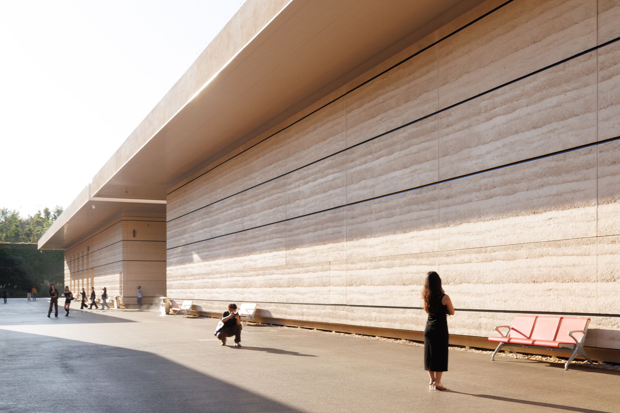 Long travertine facade with horizontal banding alongside a paved plaza with visitors and red benches in afternoon light