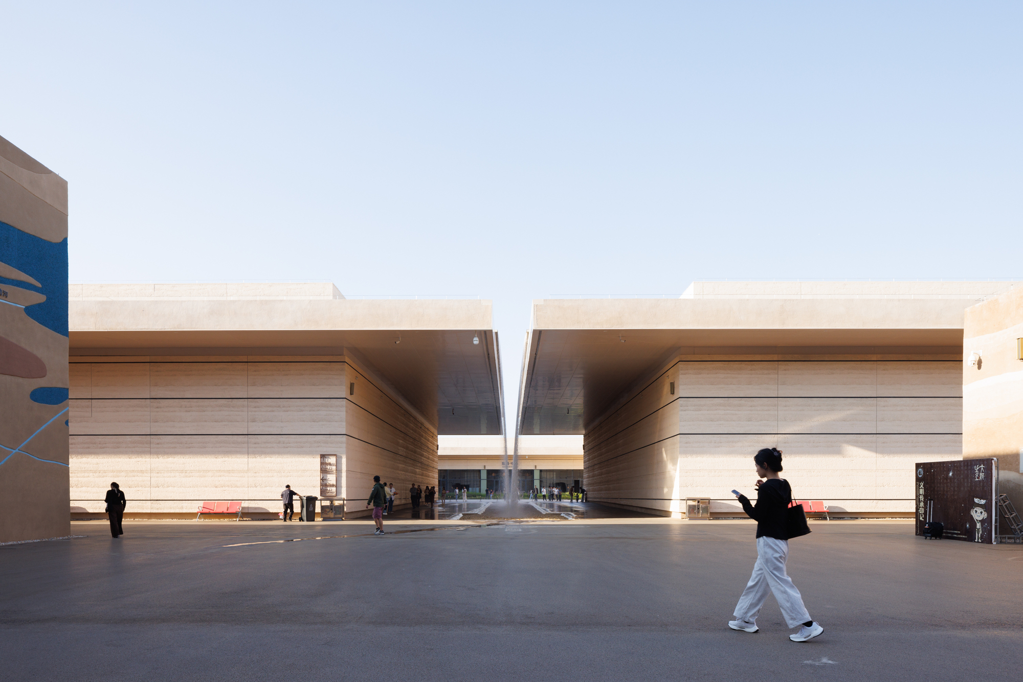 Courtyard plaza with two banded travertine volumes flanking a central slot under a pale sky